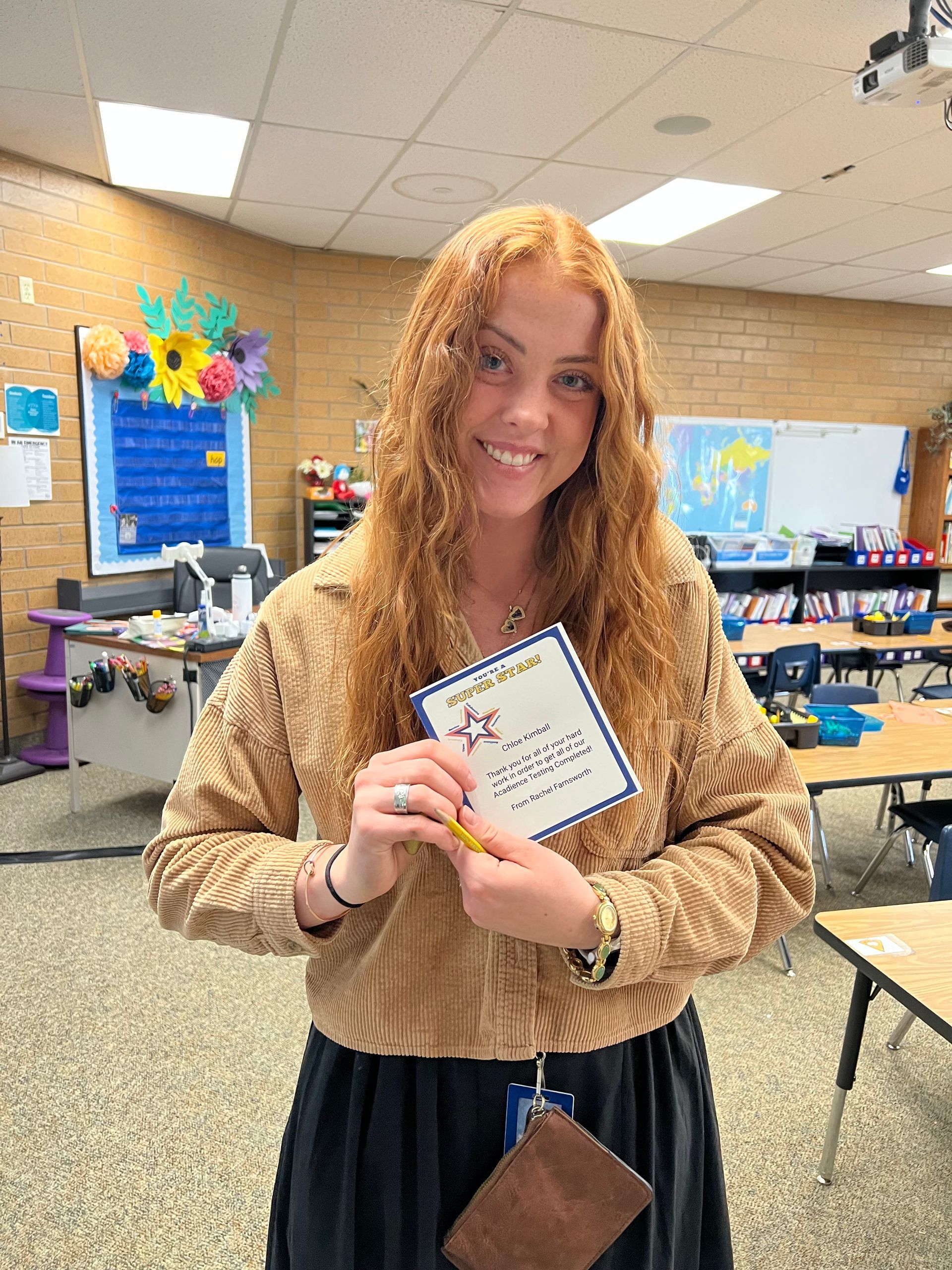 Woman with red hair smiles, holding a certificate and pencil in a classroom.