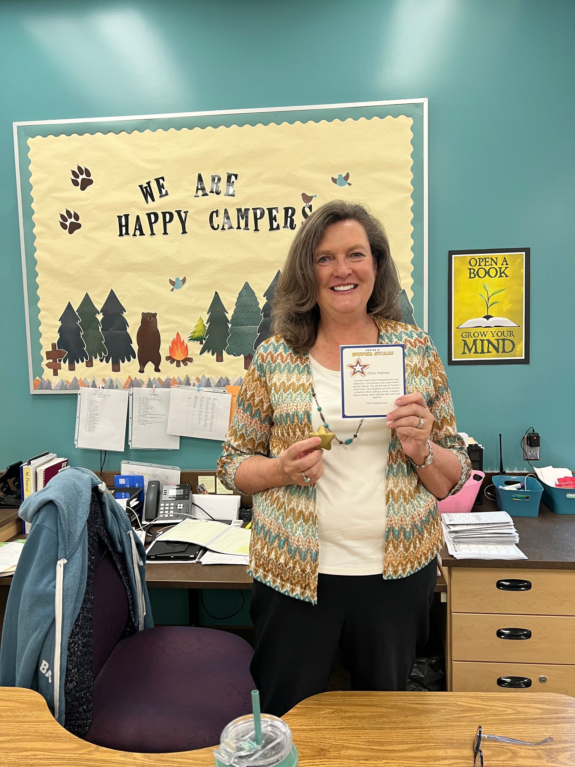 Woman in a colorful jacket holding a card and medal, smiling in front of a bulletin board that says 