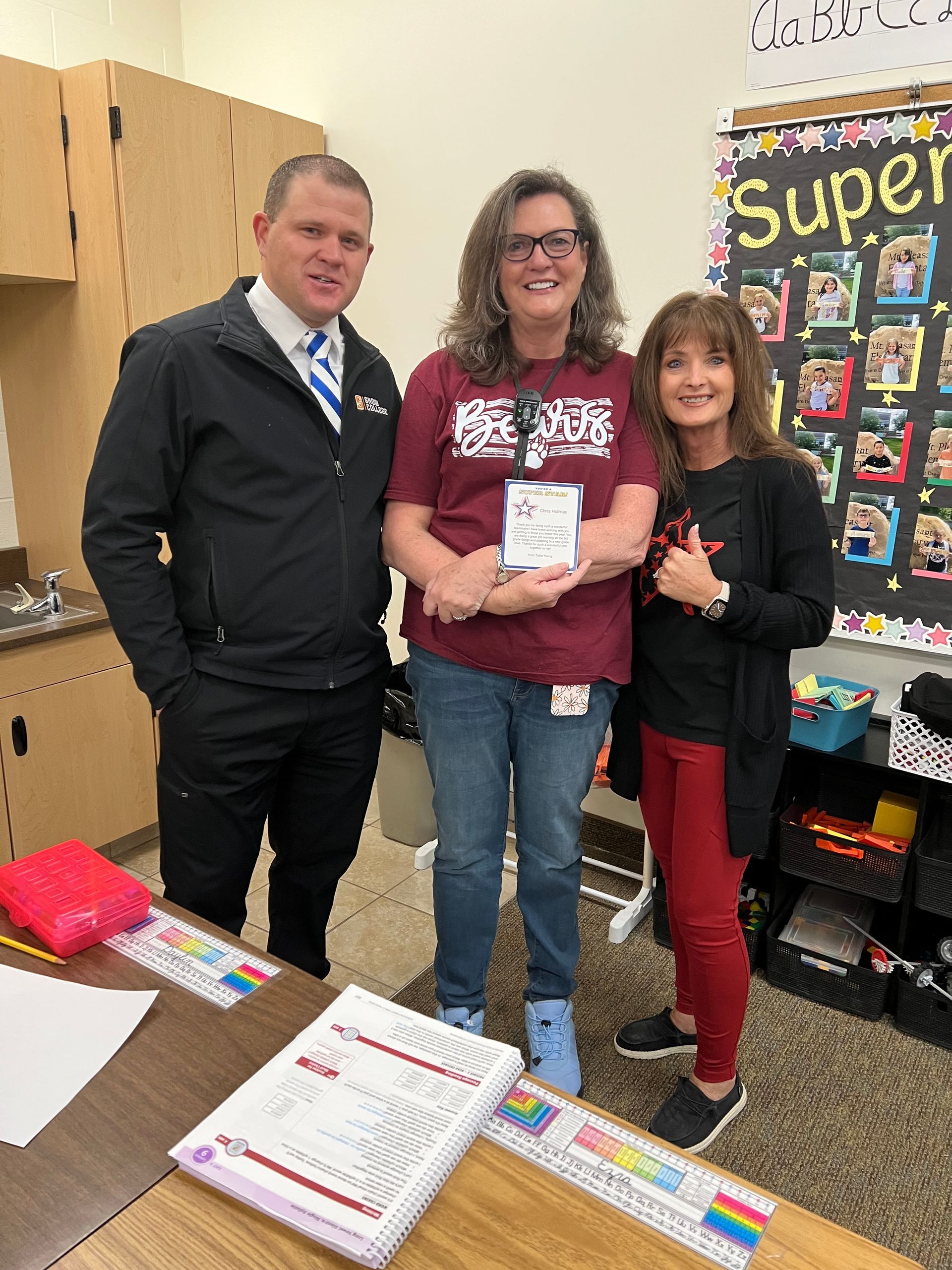 Three people in a classroom: two women and one man, posing by a desk. Woman in the center holds a certificate.