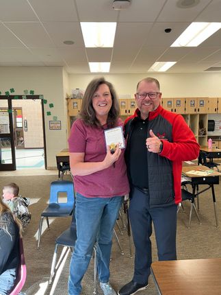Two people smiling and posing indoors, holding a small award or gift in a classroom.