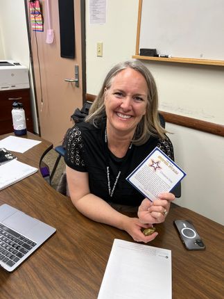 Smiling woman in office holding a card at a desk with laptop and notepad