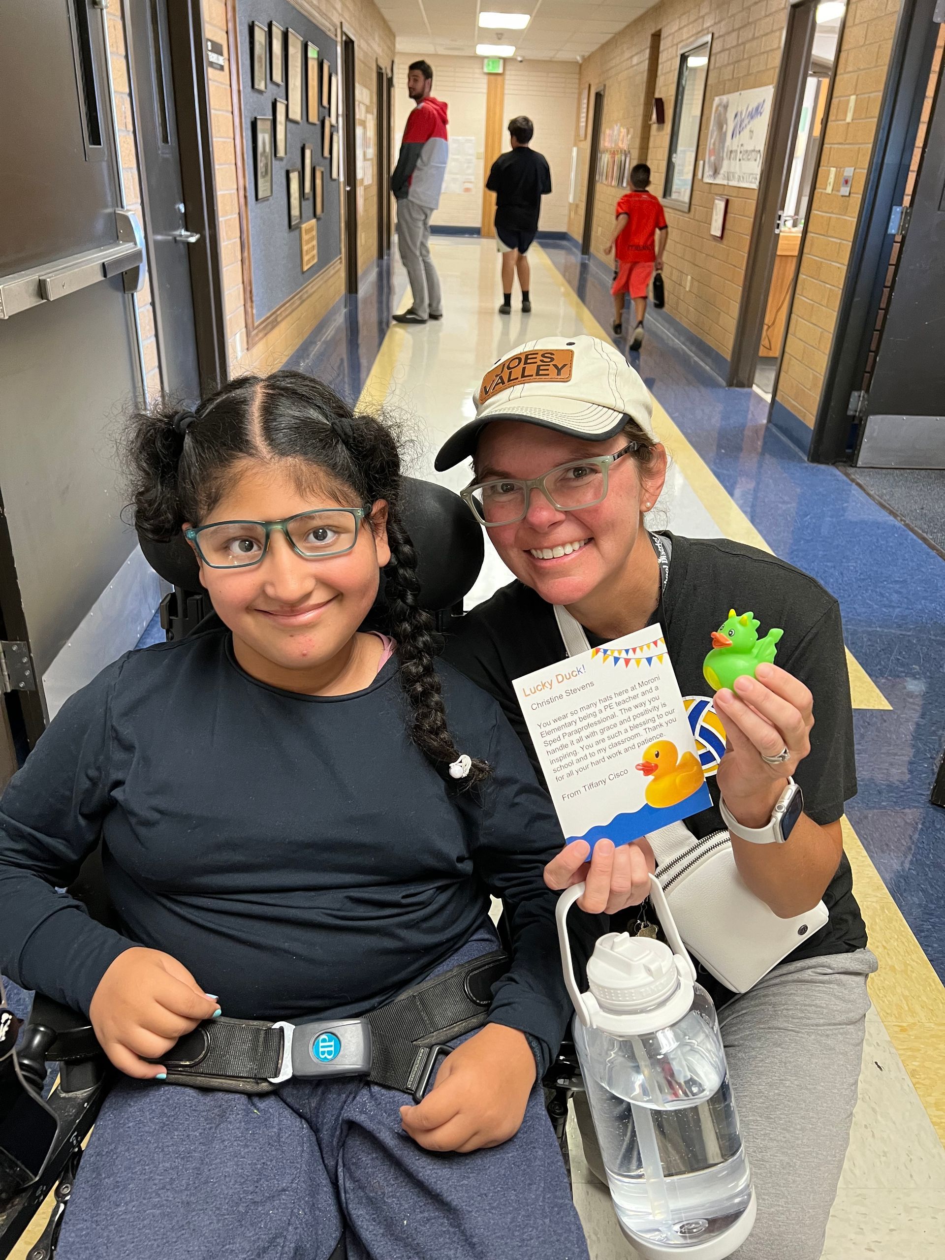 A woman is sitting next to a girl in a wheelchair in a hallway.