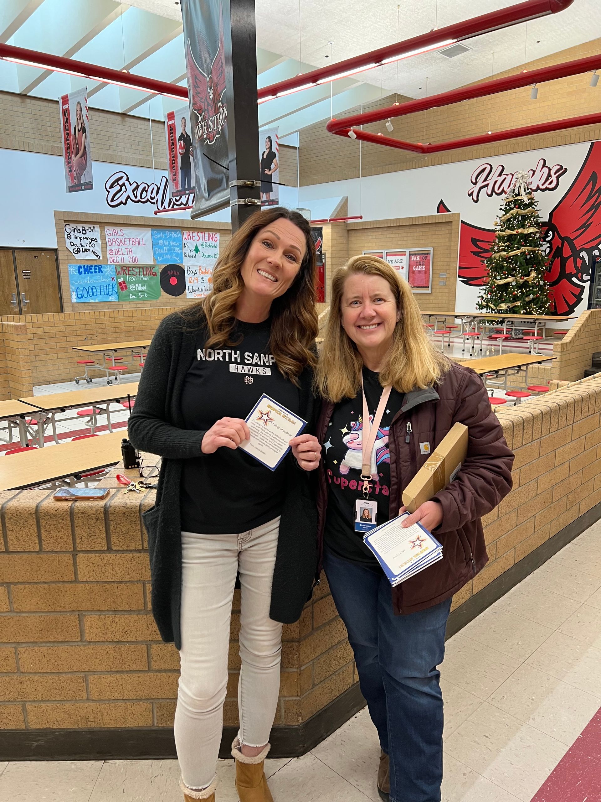 Two women in a school cafeteria holding papers and smiling. Christmas tree visible.
