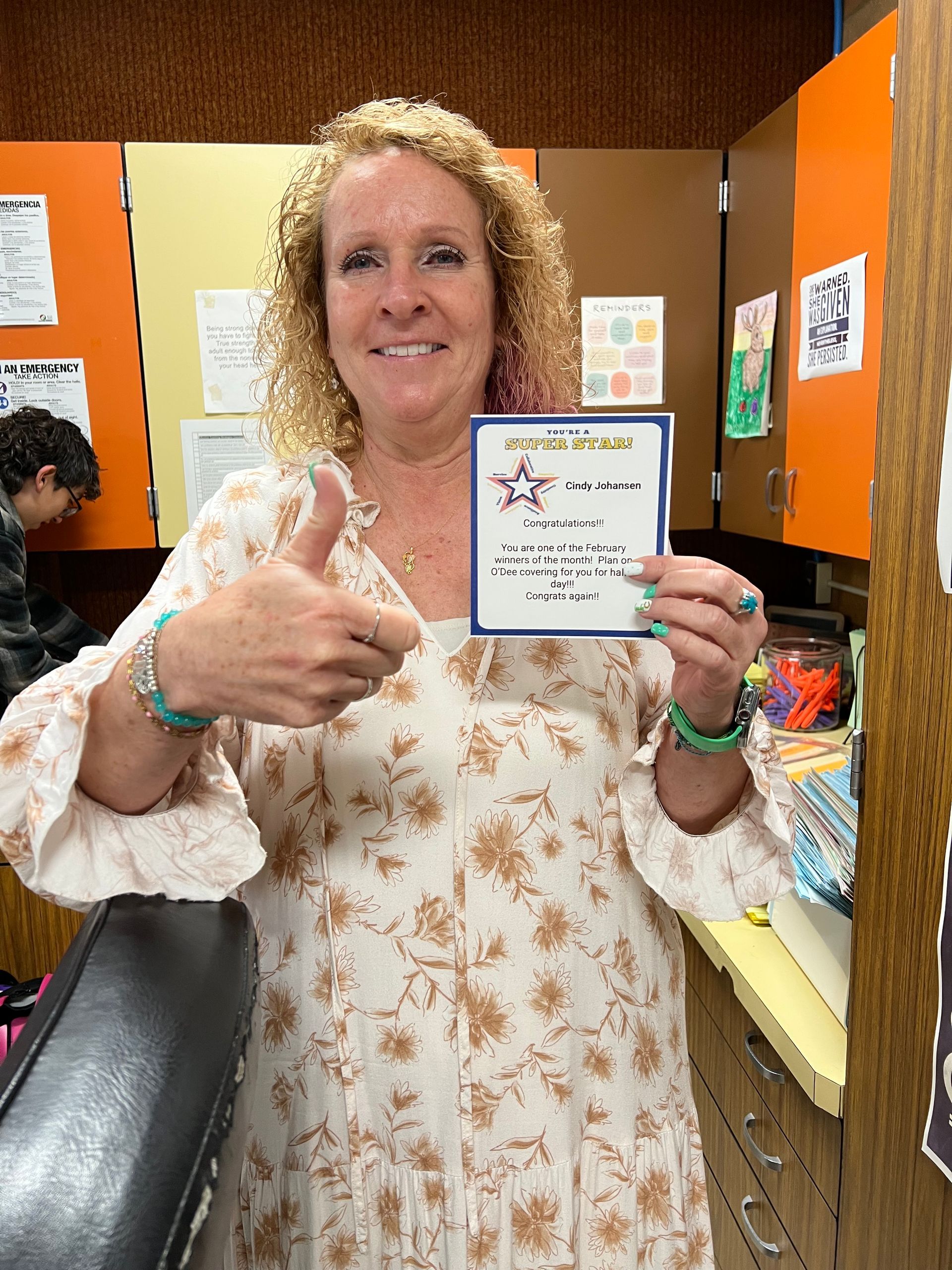 Woman smiles, gives thumbs up, holds a certificate in a room with books, orange and beige walls.