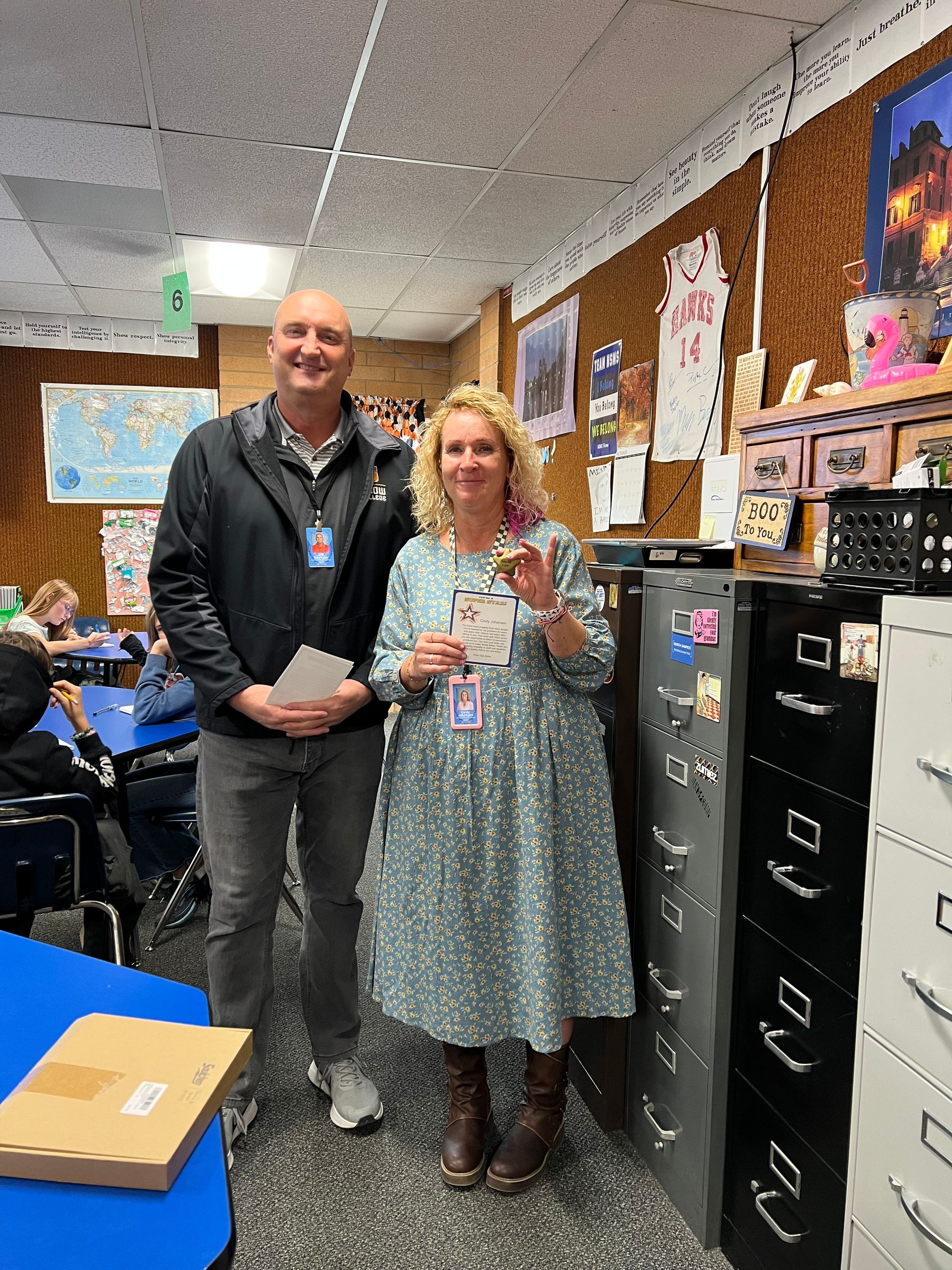 Man and woman in a classroom, the woman holding an award. They are near filing cabinets.