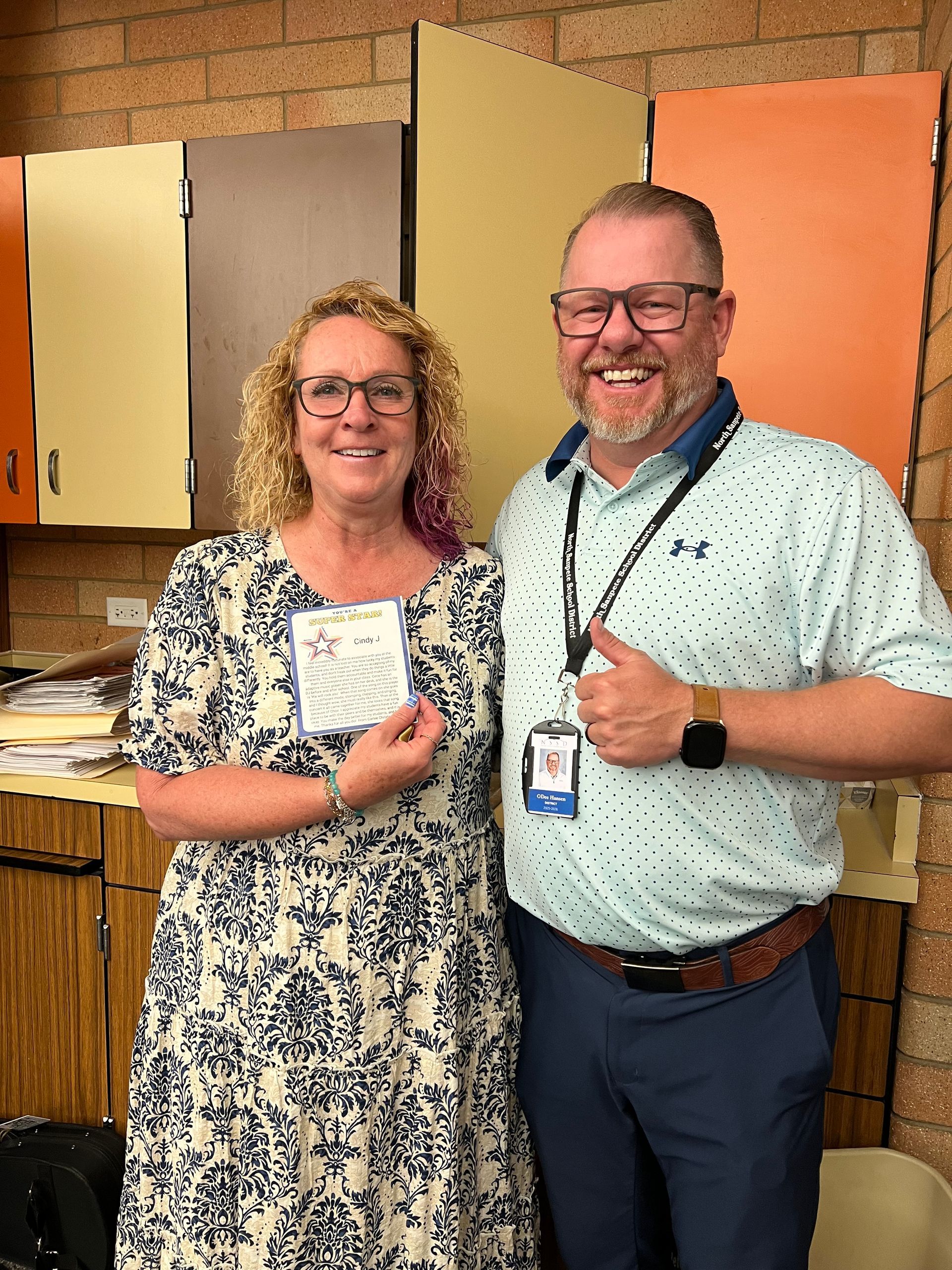 Two people smiling and holding badges in front of colorful cabinets in a classroom or office.