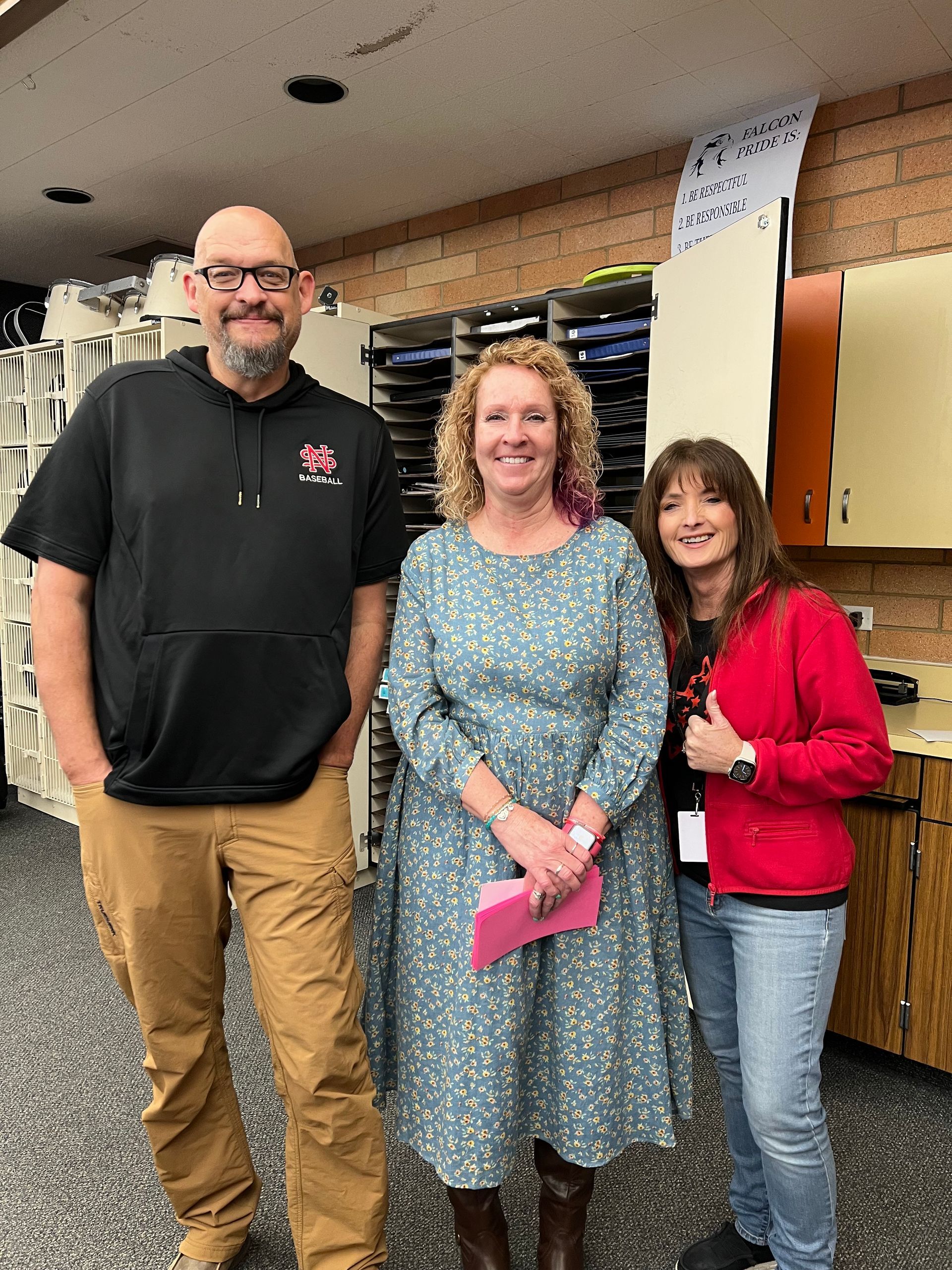 Three adults stand together in a room with shelves. One wears a black hoodie, tan pants. Another, a blue floral dress. The third, a red jacket and jeans.