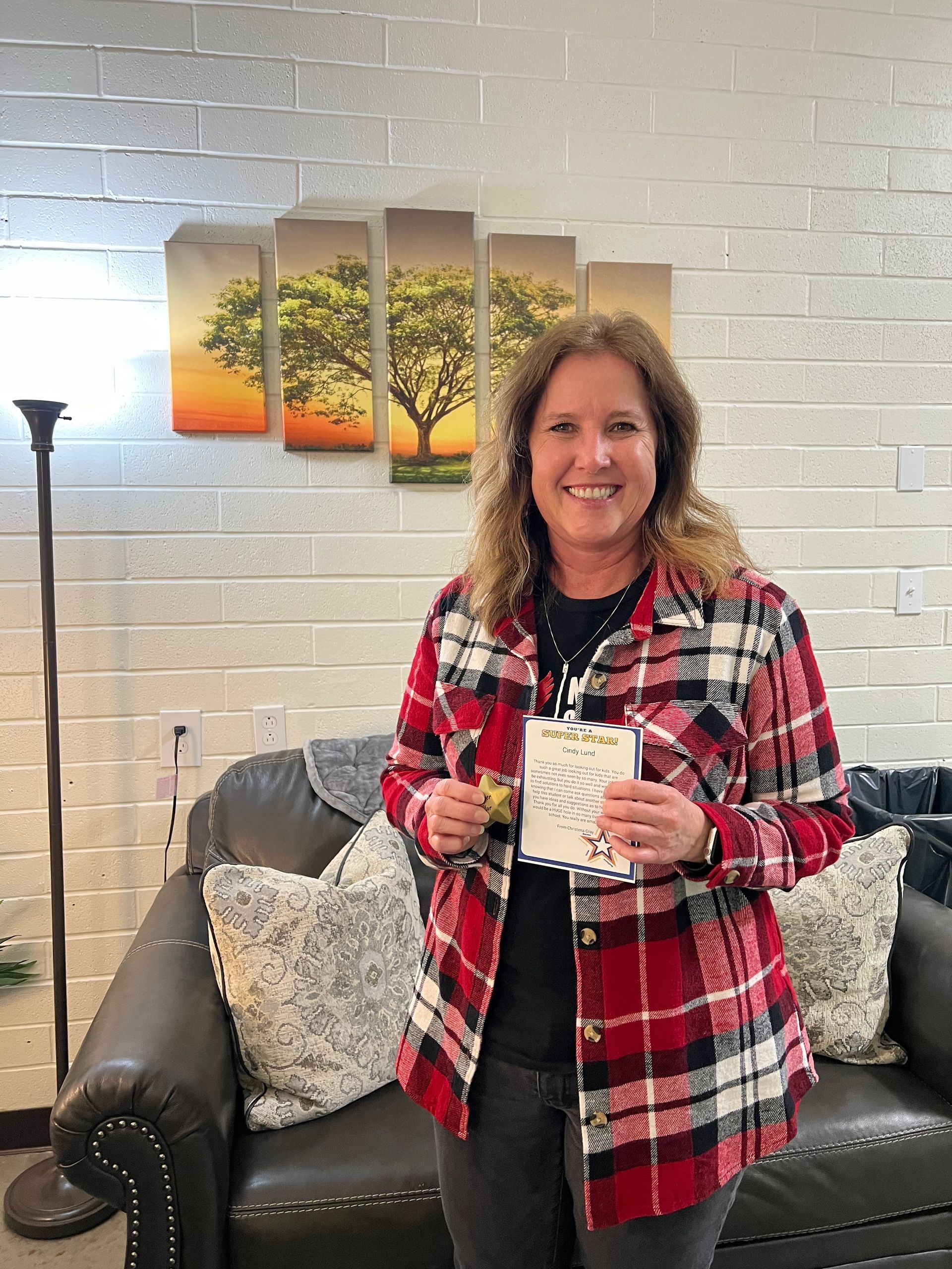 Woman in red plaid shirt smiles, holding a card. Sitting on a leather couch in a room with tree artwork.