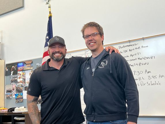 Two smiling men standing together in an office, one with an arm around the other, near a whiteboard and flag.