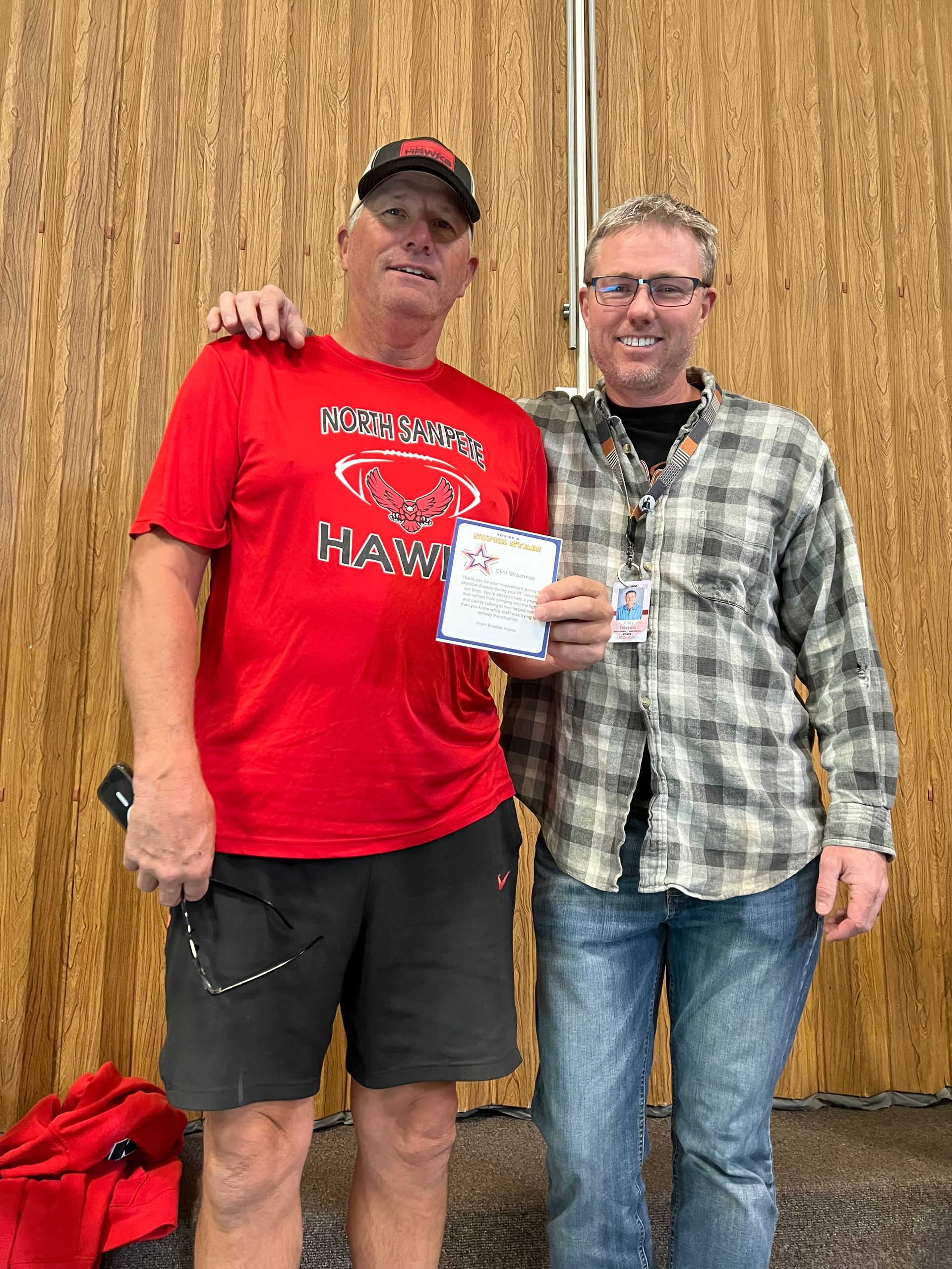 Two men pose, one in red, one in plaid holding a document. Indoors, against a wood-paneled wall.