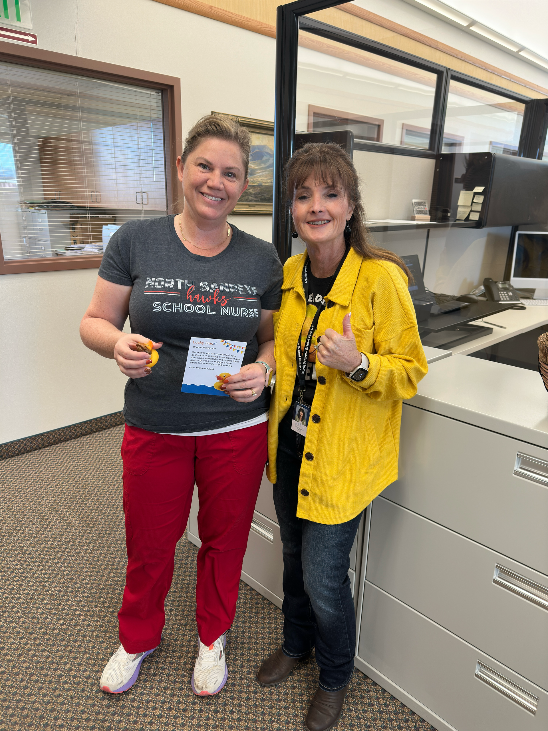 Two women are standing next to each other in an office . one of the women is wearing a yellow jacket.