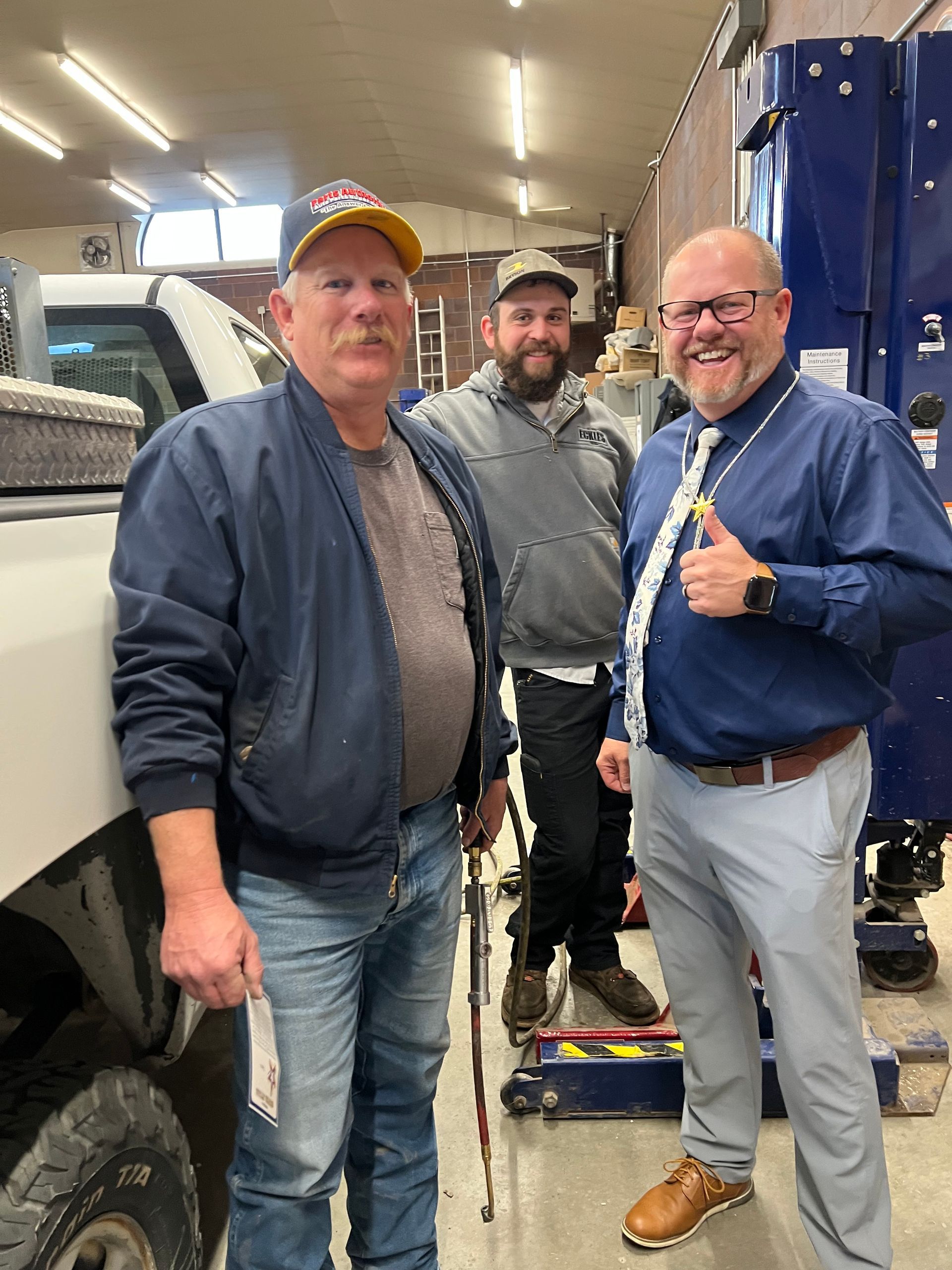 Three men pose in a garage; one holds a receipt. A man gives a thumbs-up.