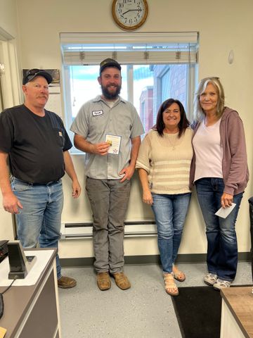 Four people standing indoors near a window. A man in the center holds a gift.