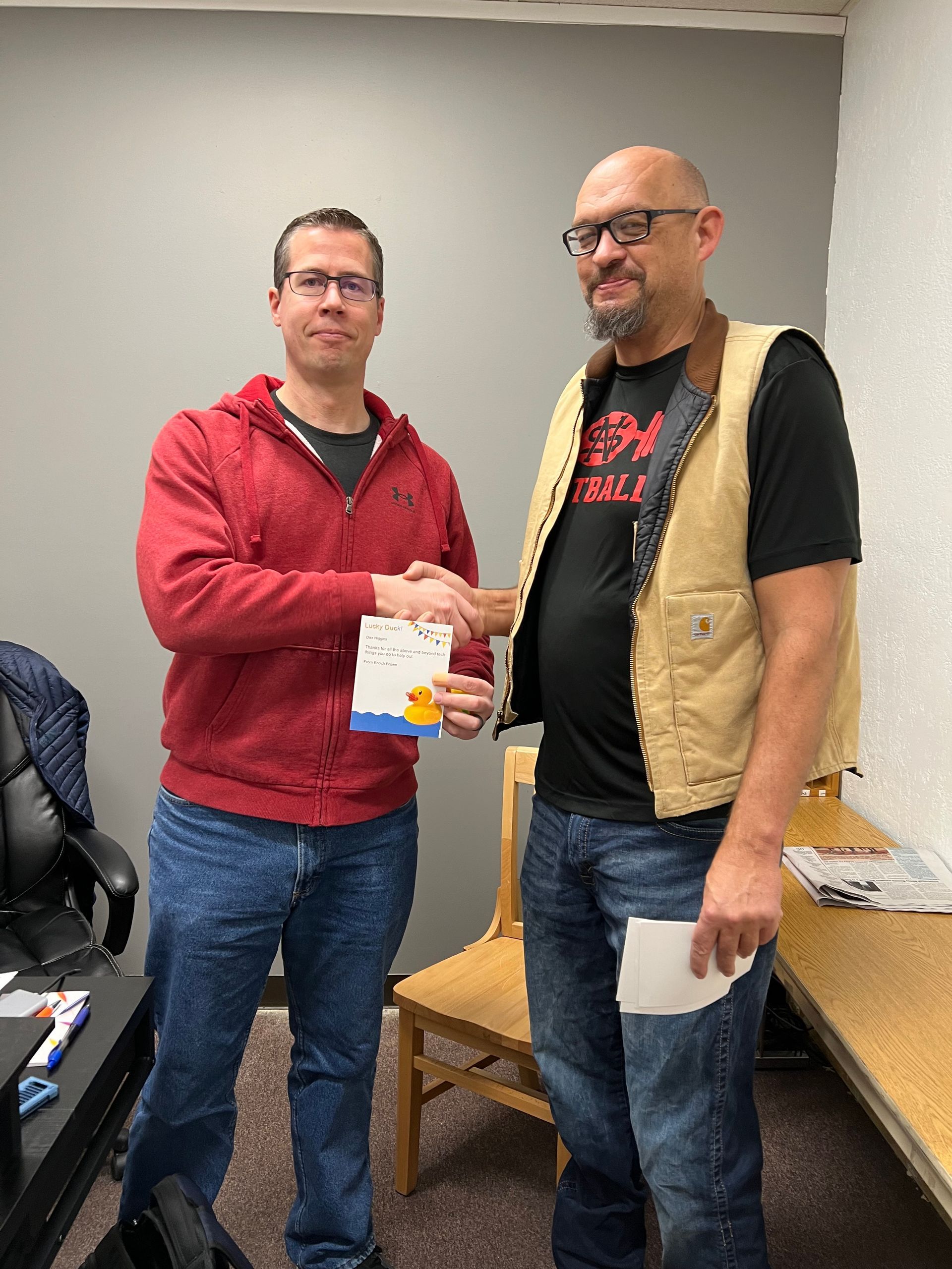 Two men are shaking hands in a room while one man is holding a piece of paper.