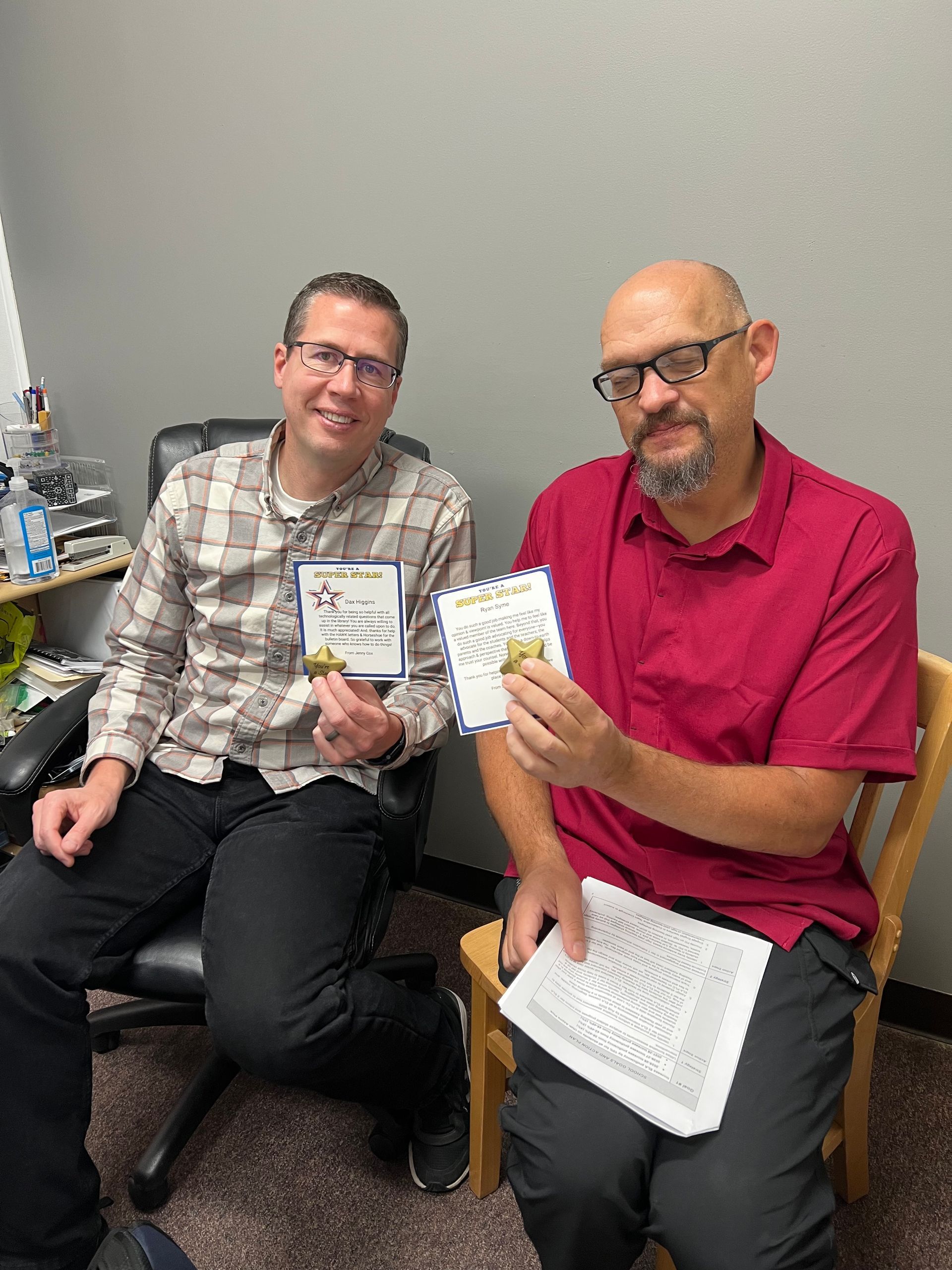 Two men in a room holding cards and objects, smiling. One in a plaid shirt, the other in red.