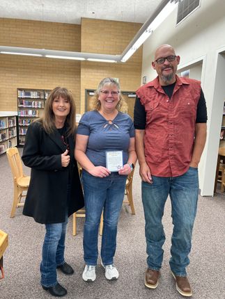 Three people standing in a library. Woman on the left in black jacket, center woman holding a paper, man on the right in a red shirt.