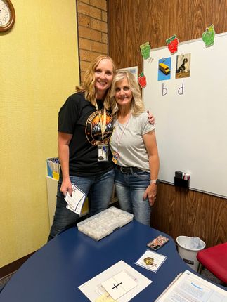 Two women smiling, arm in arm, in a classroom. One wears a black shirt, the other a grey top and jeans.