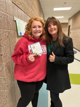 Two women posing in a hallway. One in a red hoodie holds an award; the other in a black coat gives a thumbs up.
