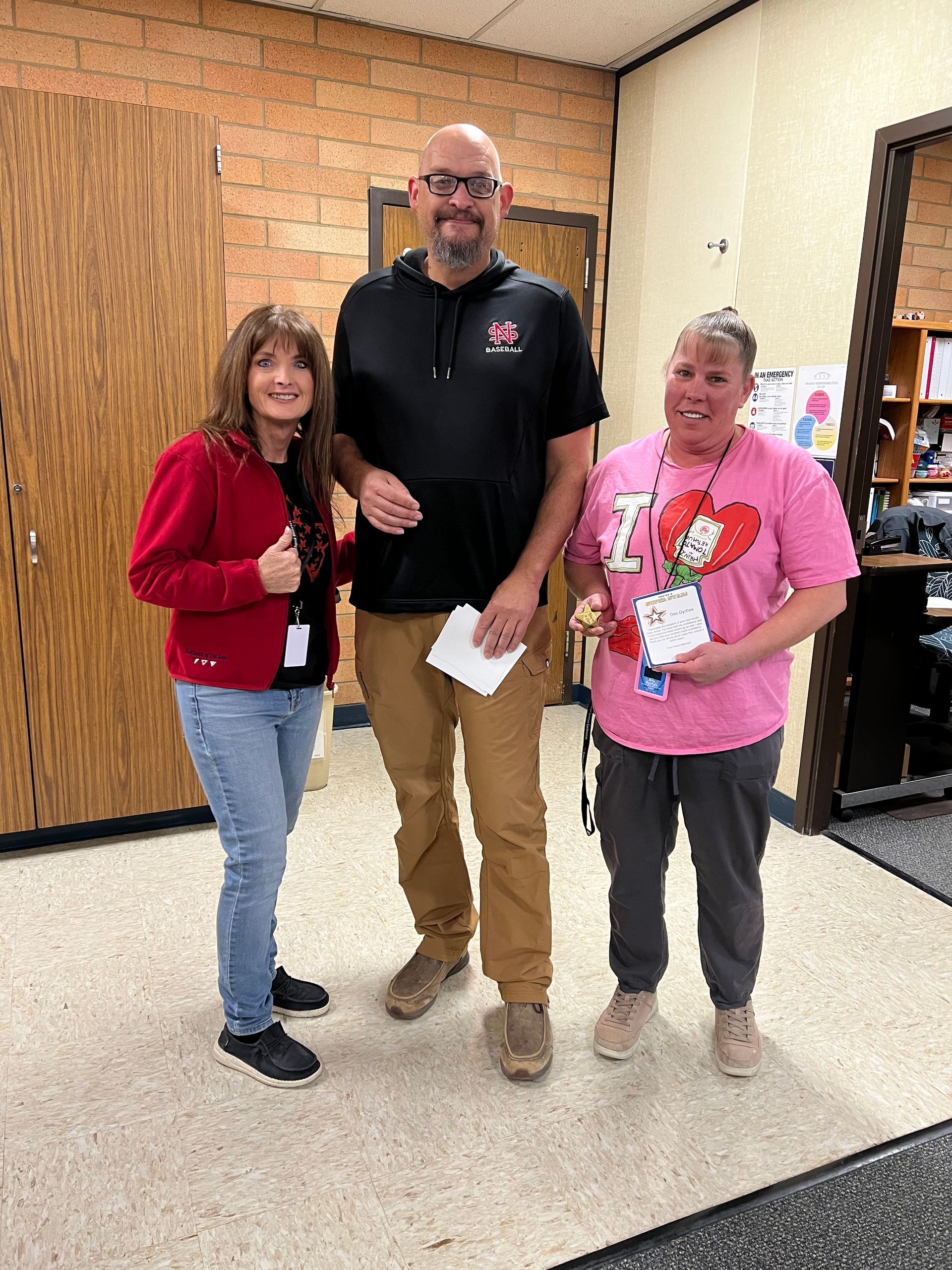 Three people standing in a hallway. One in red, one in black, and one in pink. They are holding papers.
