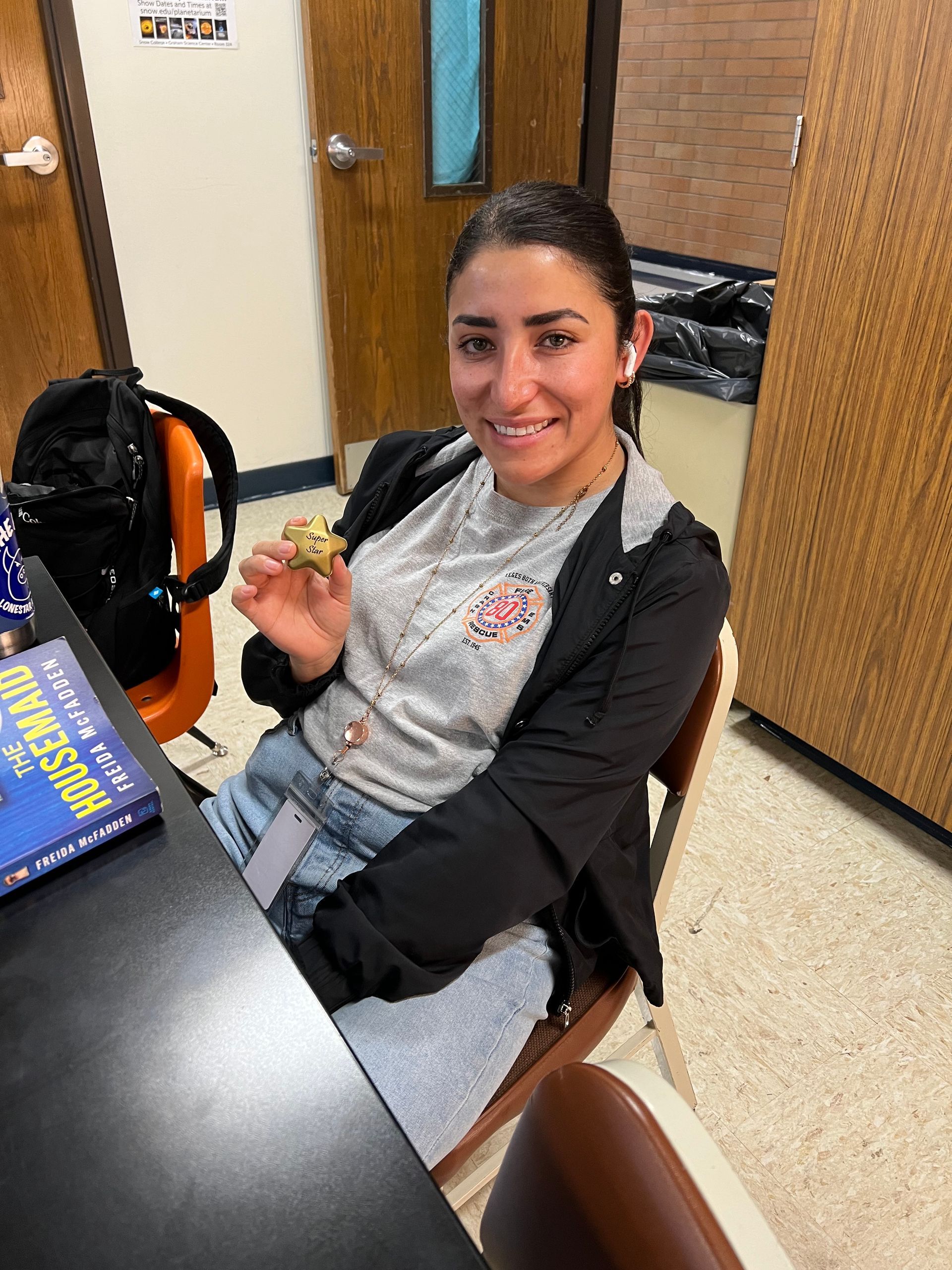 Person smiling at a desk in an office, holding a small snack and wearing a black jacket and gray outfit.