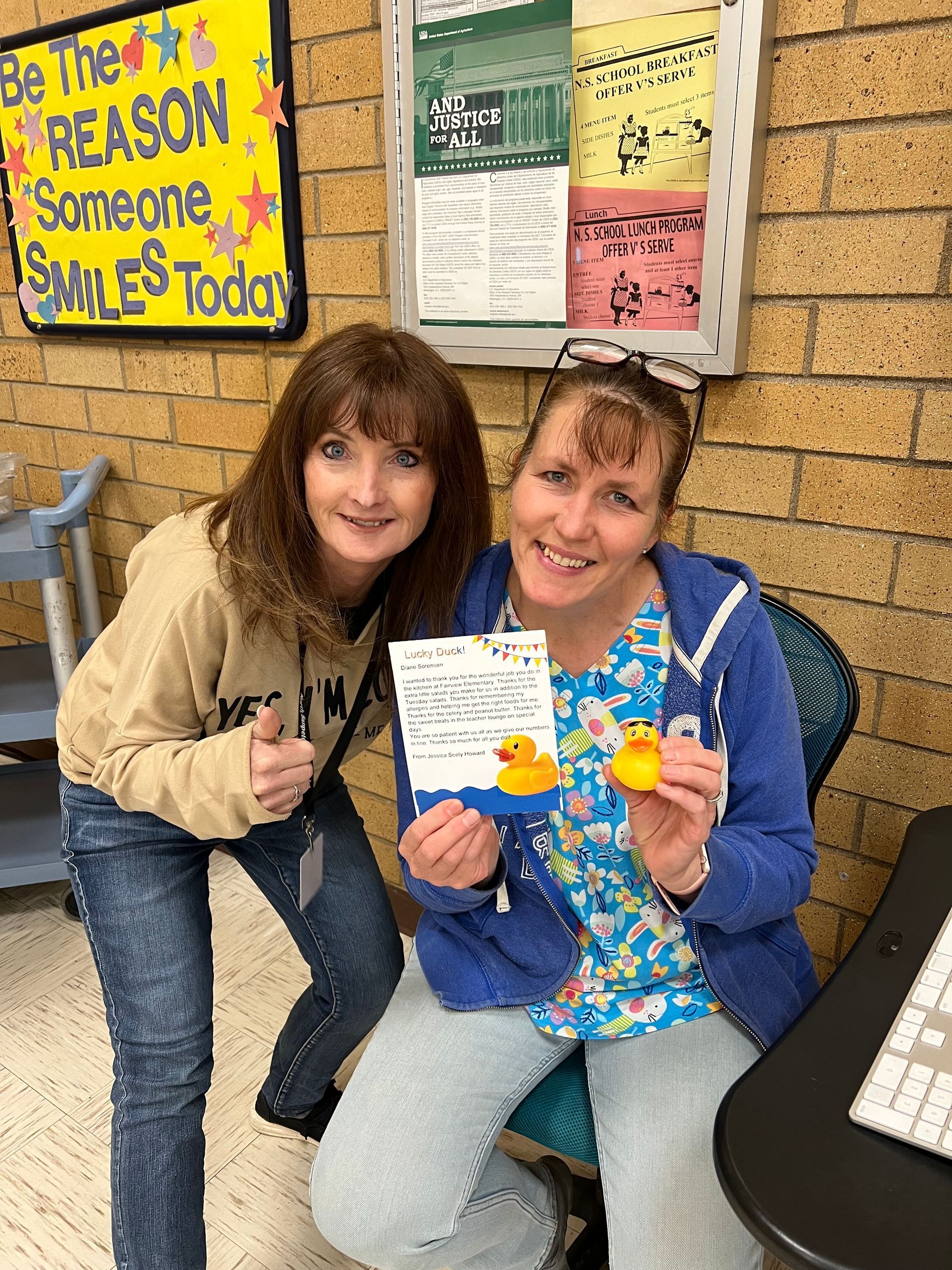 Two women are posing for a picture in front of a sign that says `` be the reason someone smiles today ''.