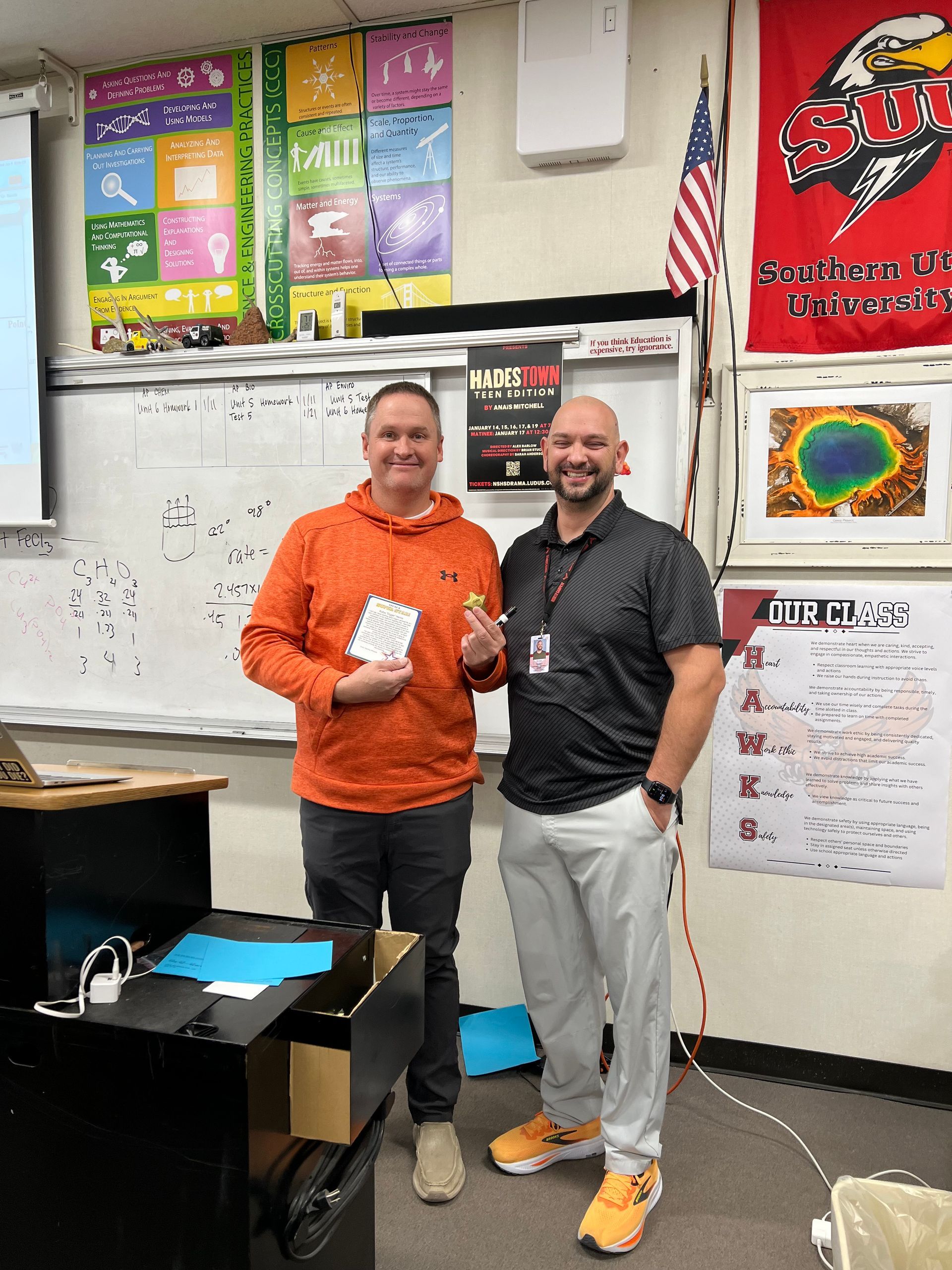 Two men smiling, holding items in a classroom with whiteboard, flag, posters, and equipment.
