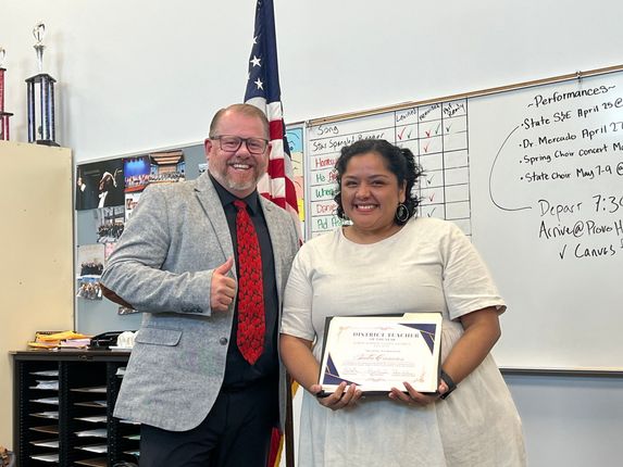 Two people smile, holding a certificate in a classroom with a whiteboard and American flag.