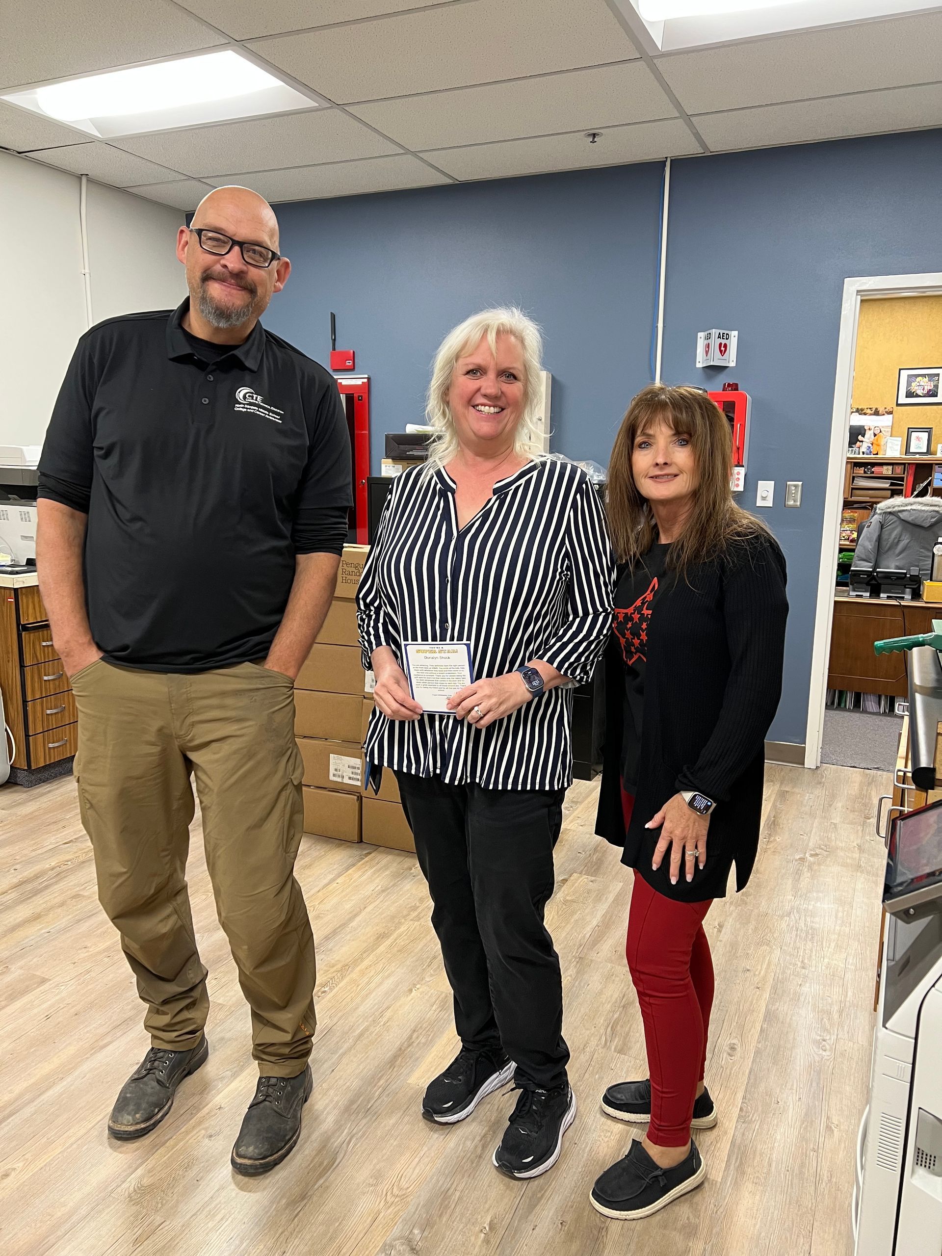 Three people smiling indoors; woman in striped shirt holds a check, others stand nearby.
