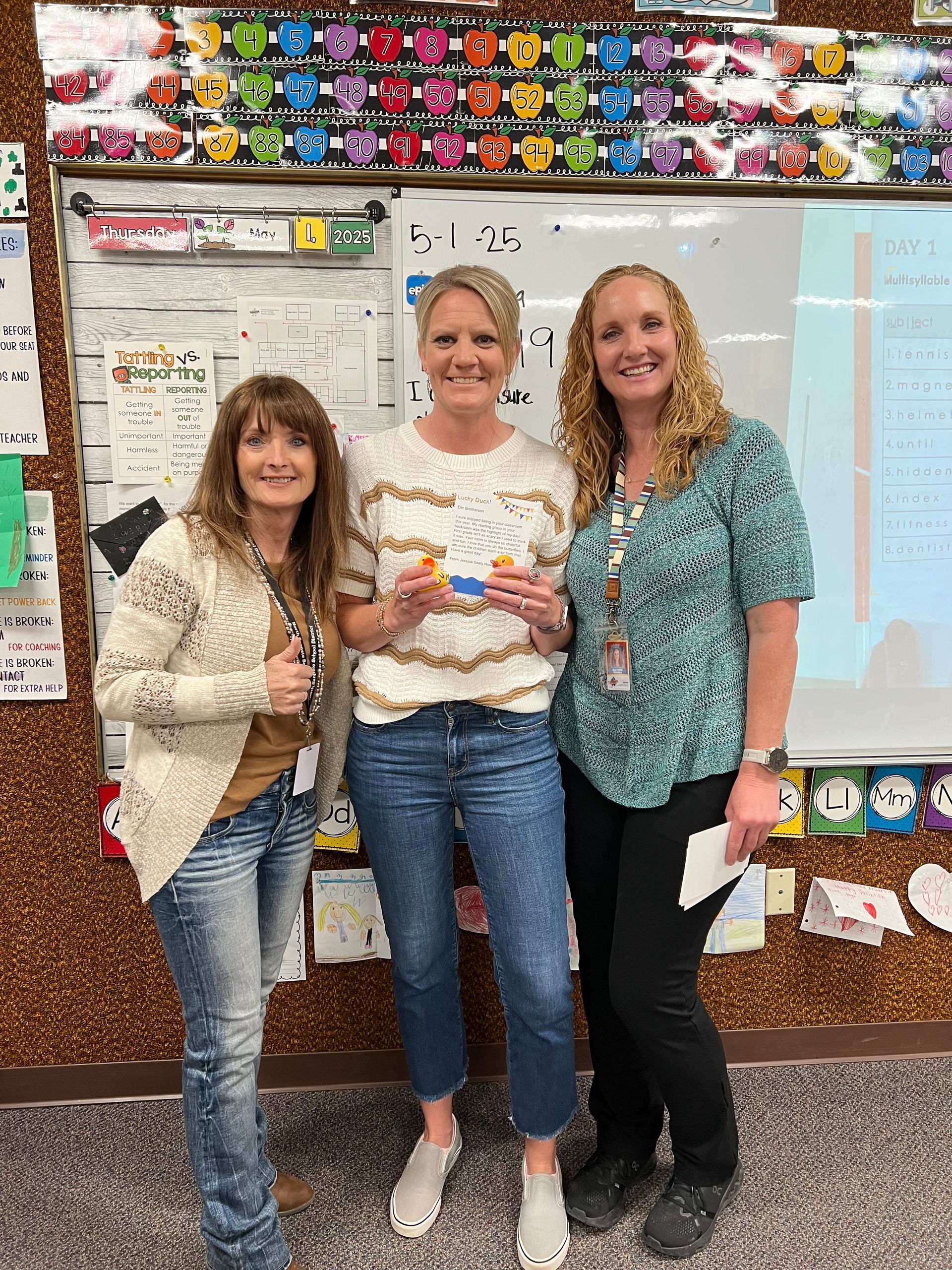 Three women are standing next to each other in front of a whiteboard in a classroom.