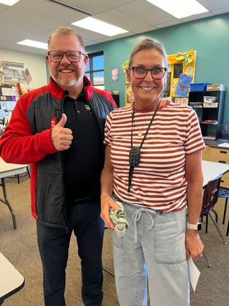 Two smiling people standing indoors, one giving a thumbs-up in a classroom.