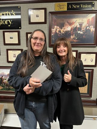 Two women standing in front of a wall with framed documents and artwork; one holding a clipboard, the other giving a thumbs-up.