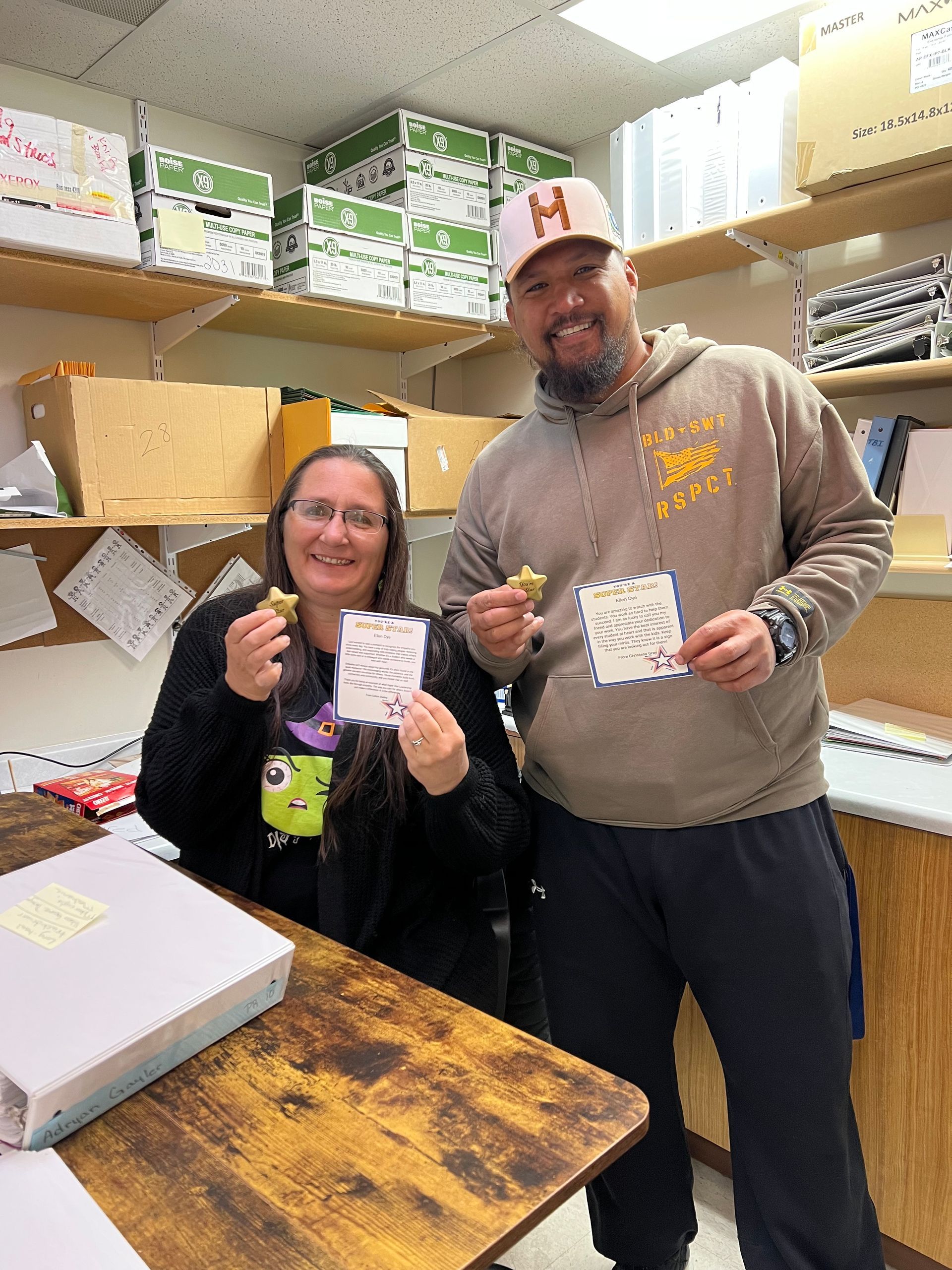 Two people holding star-shaped items and cards, smiling in an office with storage shelves and boxes.