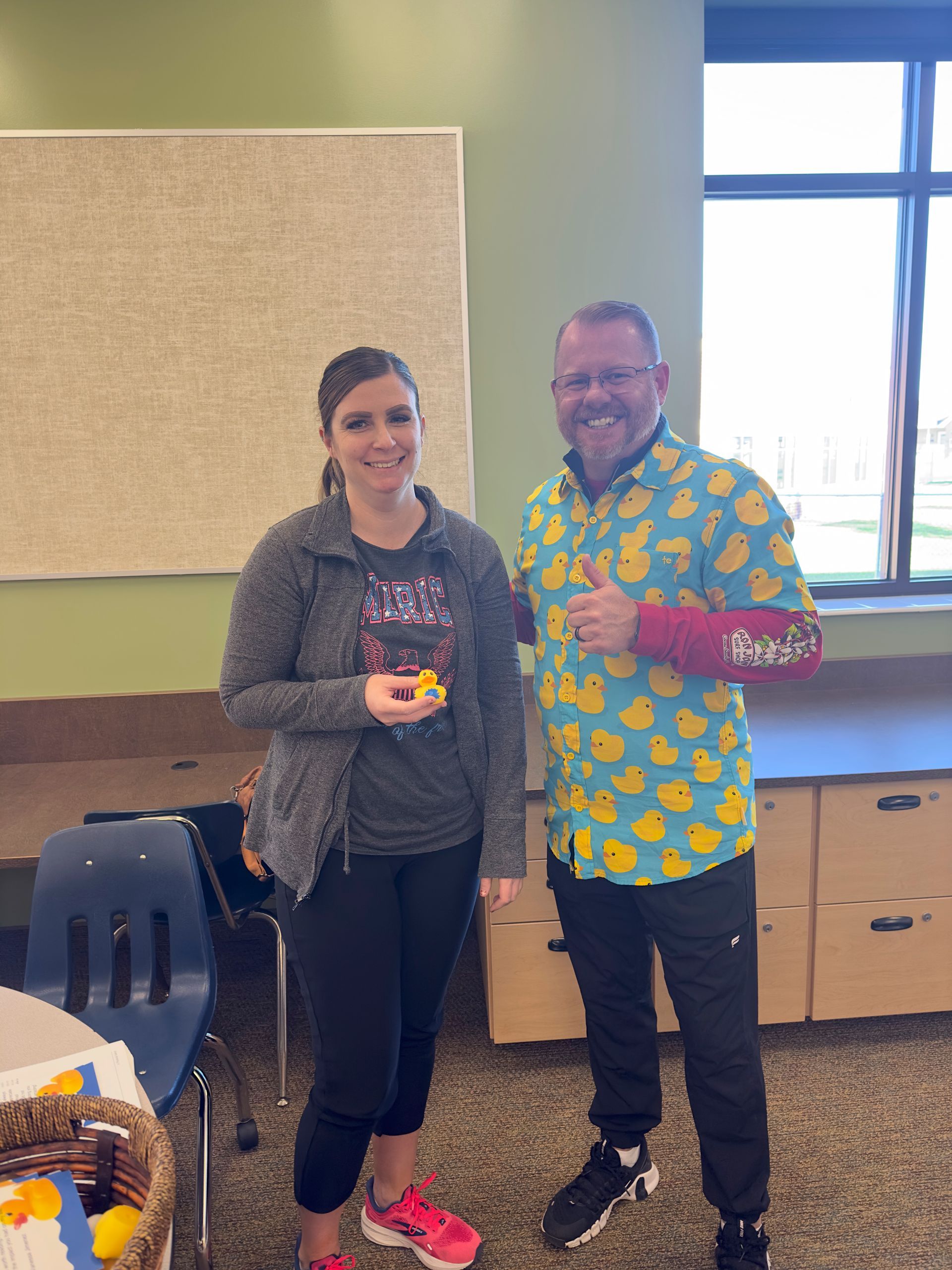 A man and a woman are standing next to each other in a room . the man is wearing a duck shirt.