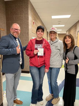 Four people in a school hallway: a man giving a thumbs-up, a woman holding a paper, and two other women.