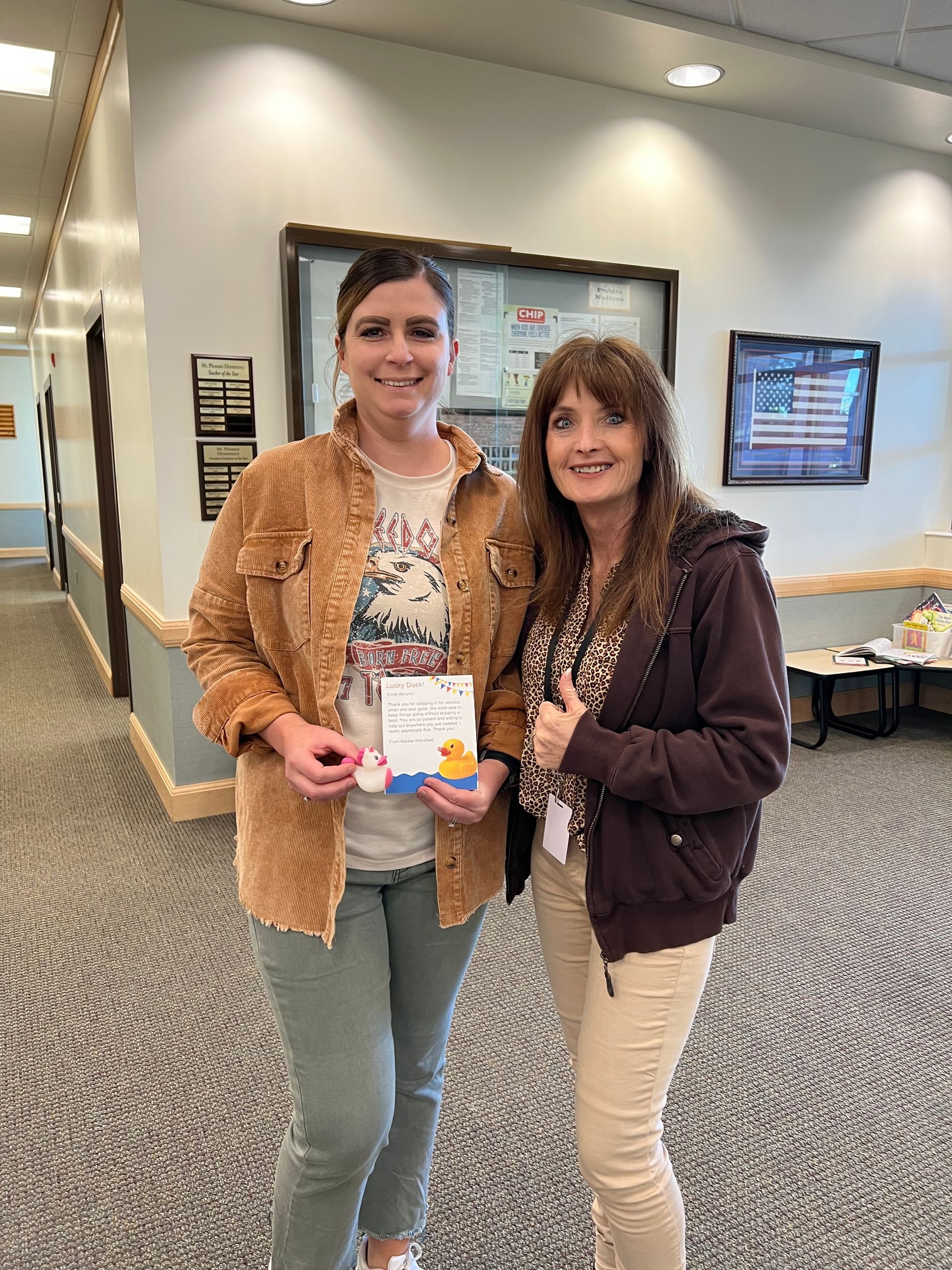 Two women are standing next to each other in a hallway holding a book.
