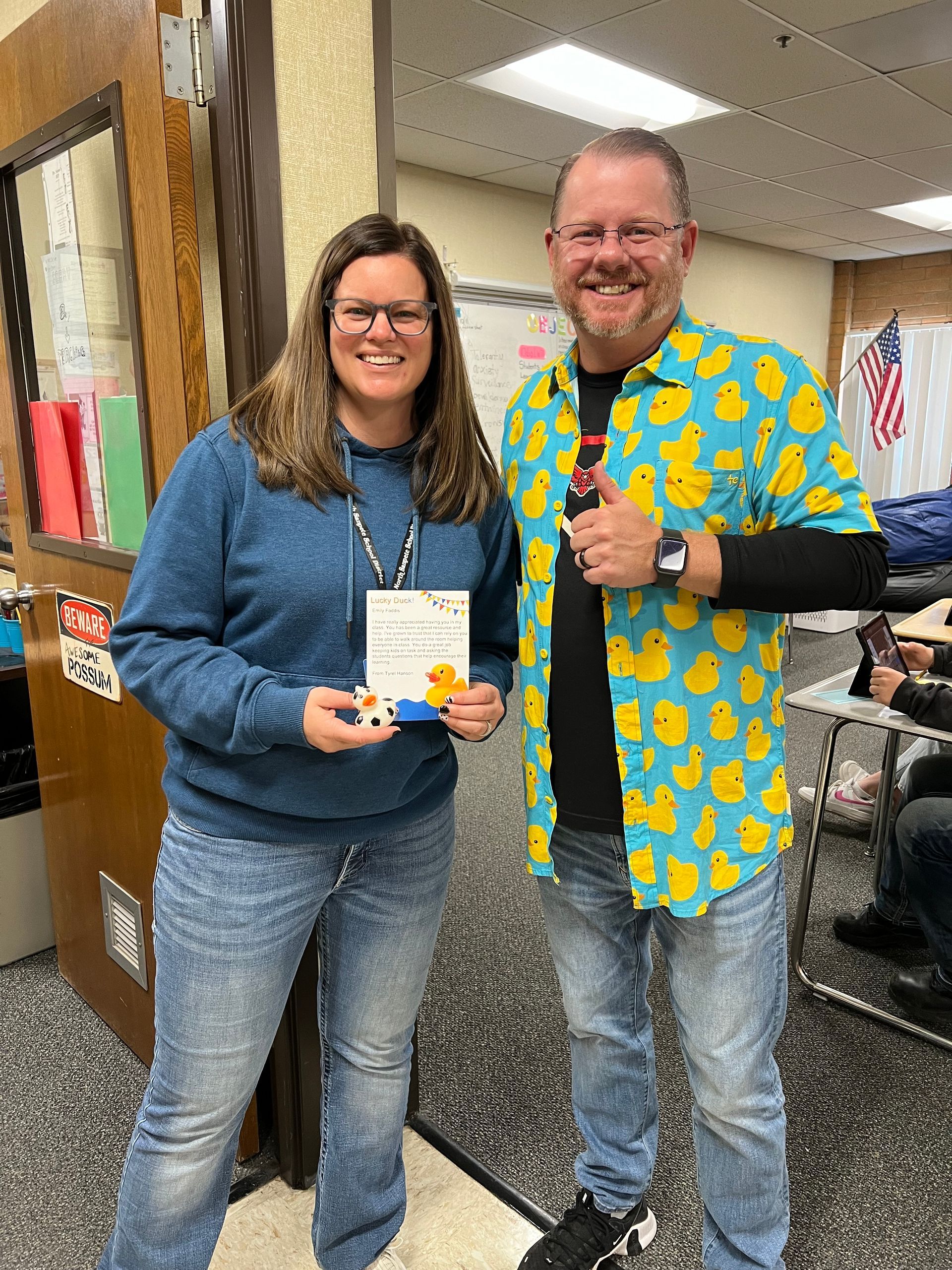 A man and a woman are standing next to each other in a room . the man is wearing a duck shirt.