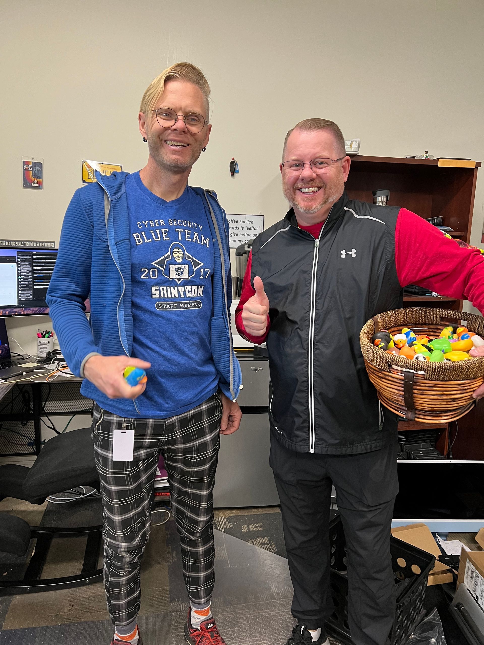 Two men are standing next to each other in an office holding a basket of candy.