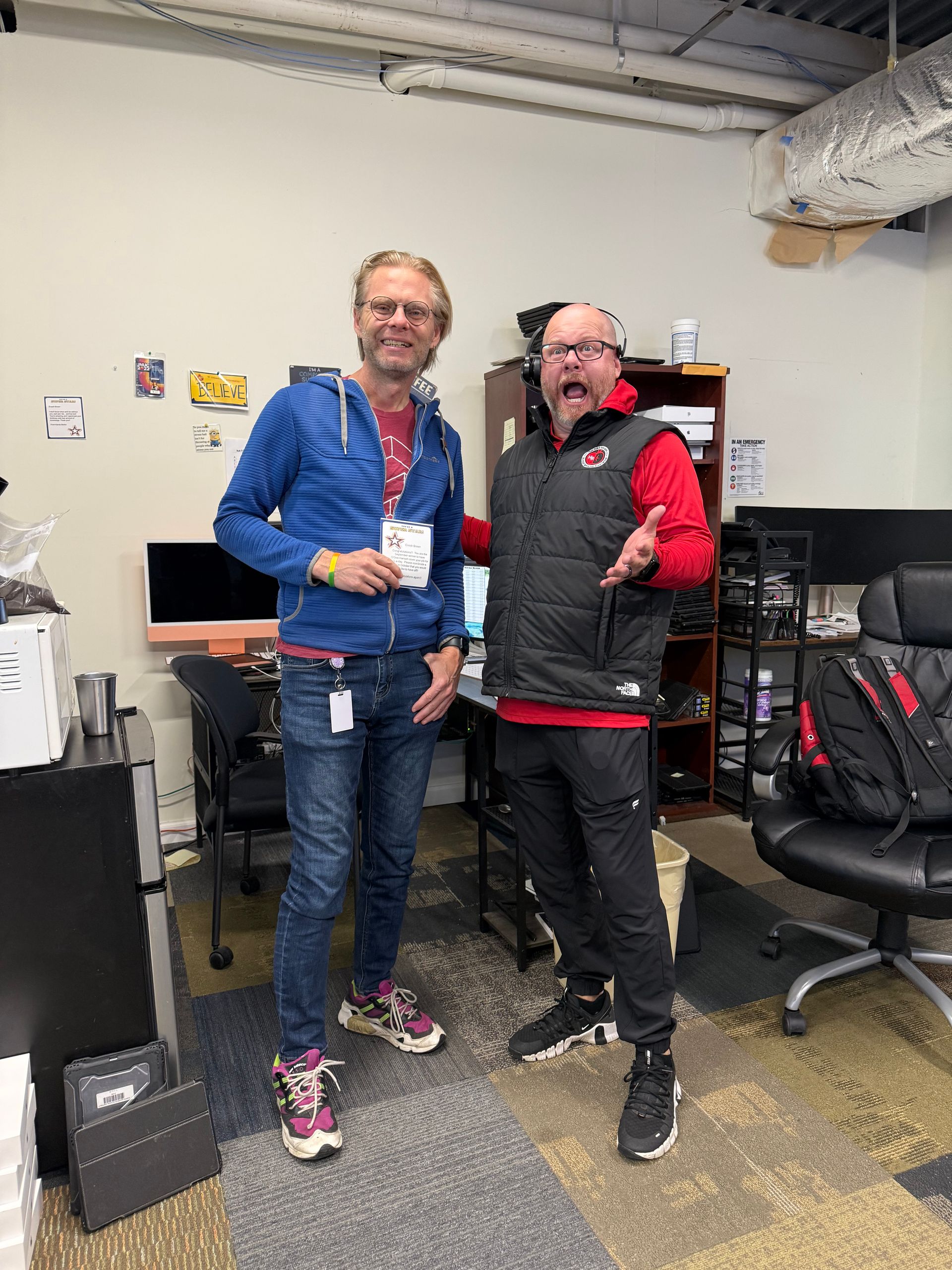 Two men stand in an office. One wears a blue jacket and jeans. The other wears a black vest and pants, gesturing.