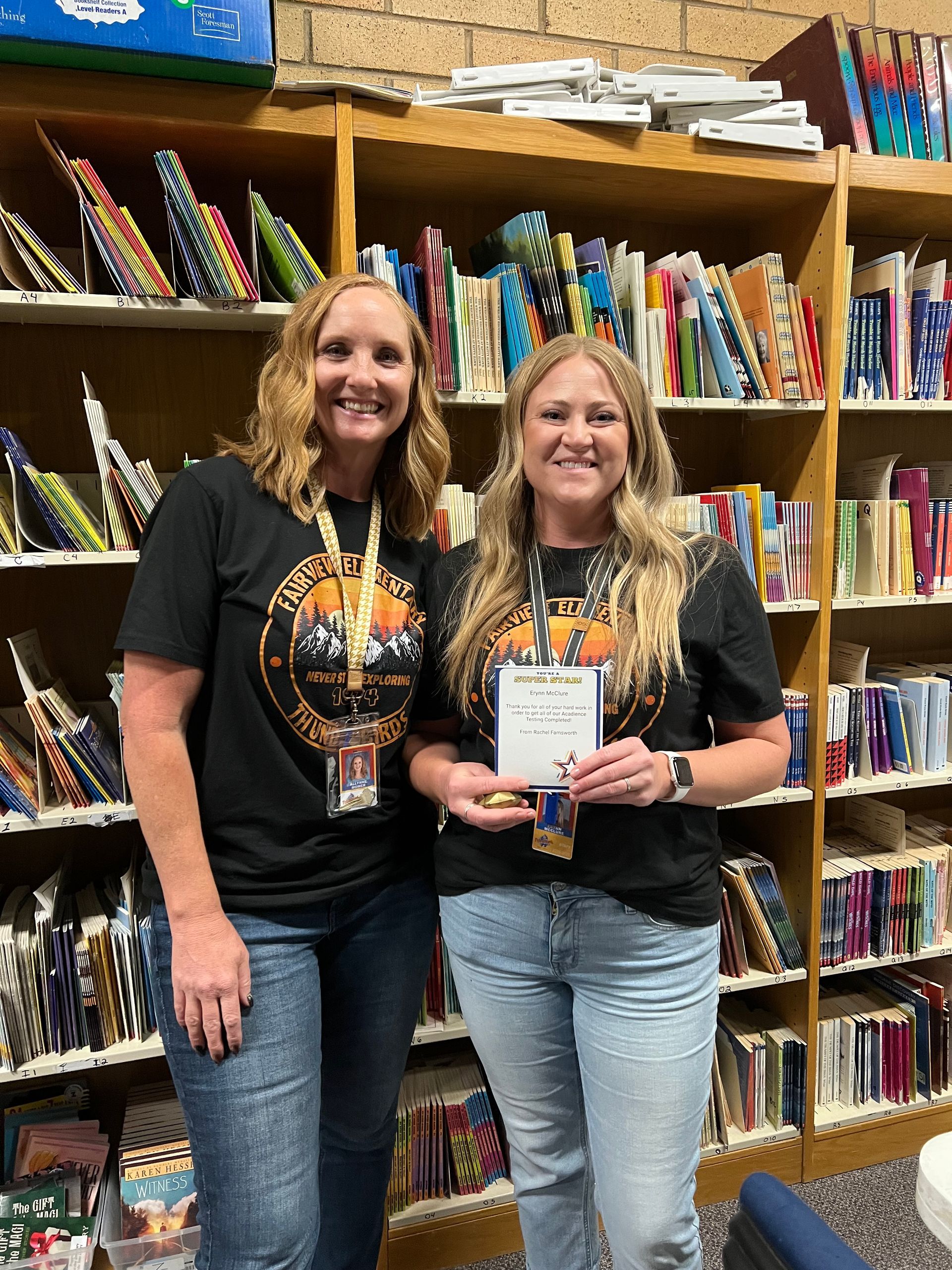 Two women pose in front of a bookshelf, one holding an award. Both wear matching black shirts, jeans, and lanyards.