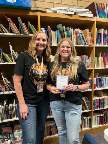 Two women pose in a library, one holding an award. Both wear black shirts and jeans.