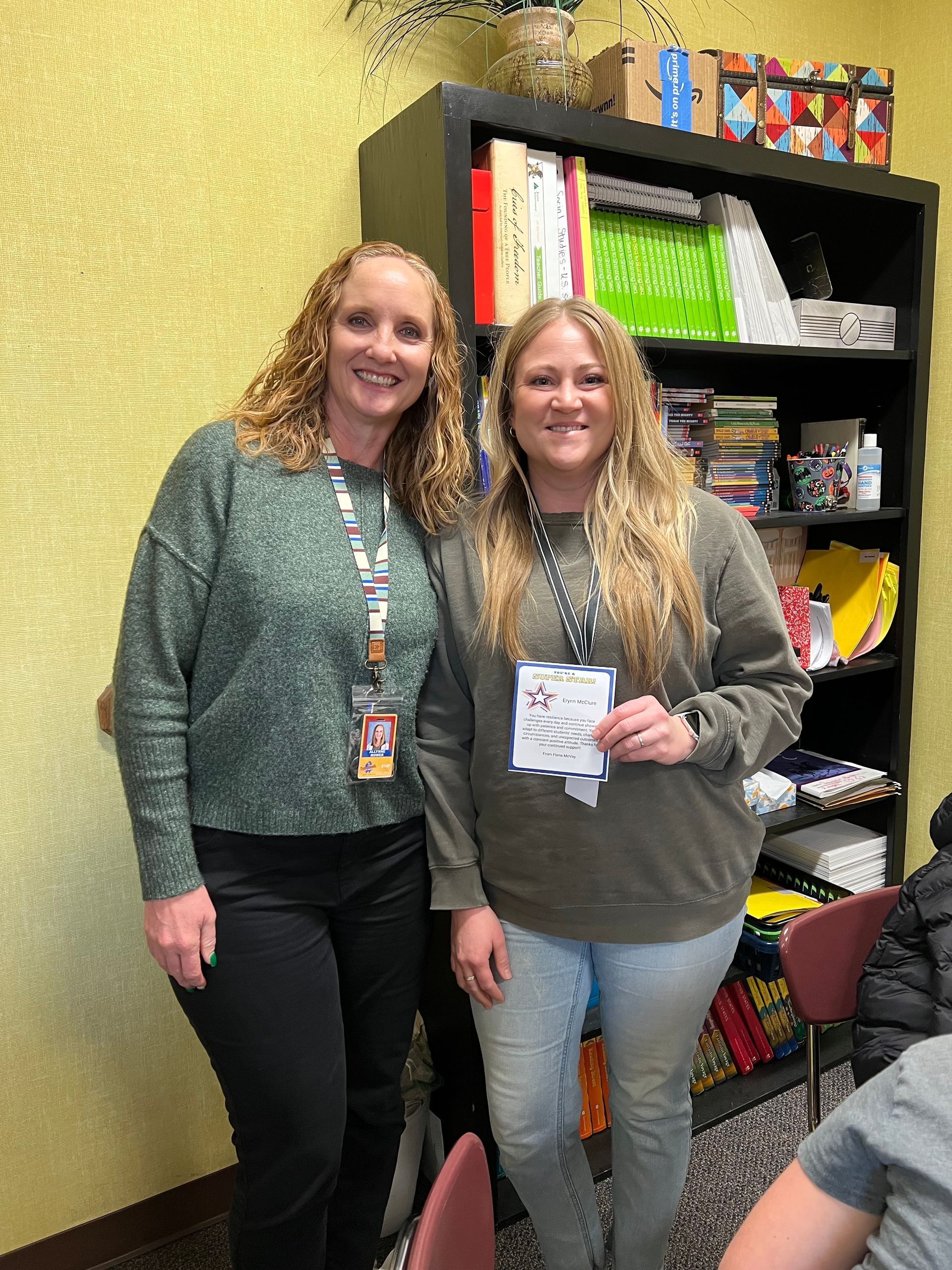 Two women stand near a bookshelf. One holds a paper. They smile in an office setting.