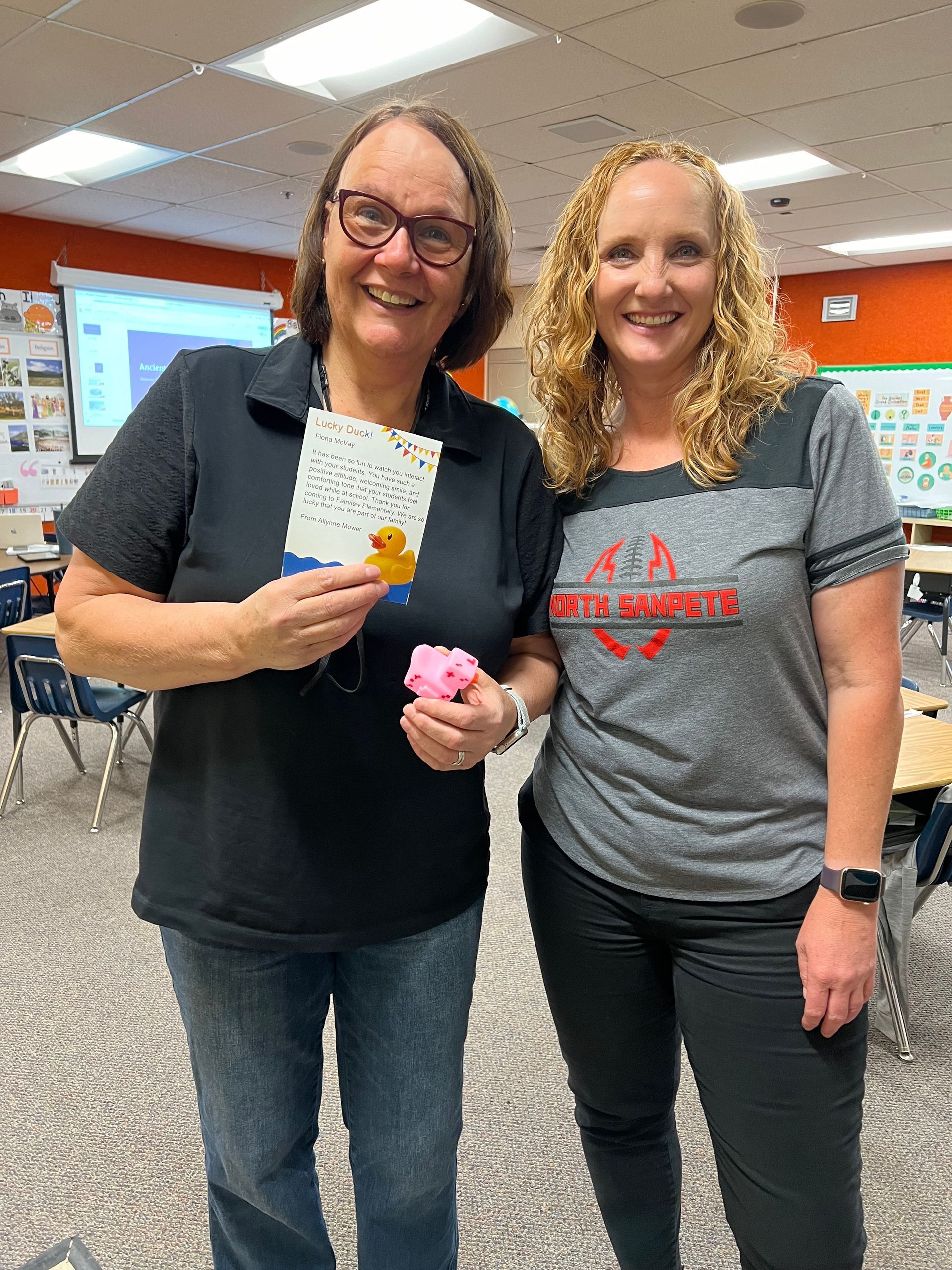 Two women are standing next to each other in a classroom holding a card and a flower.