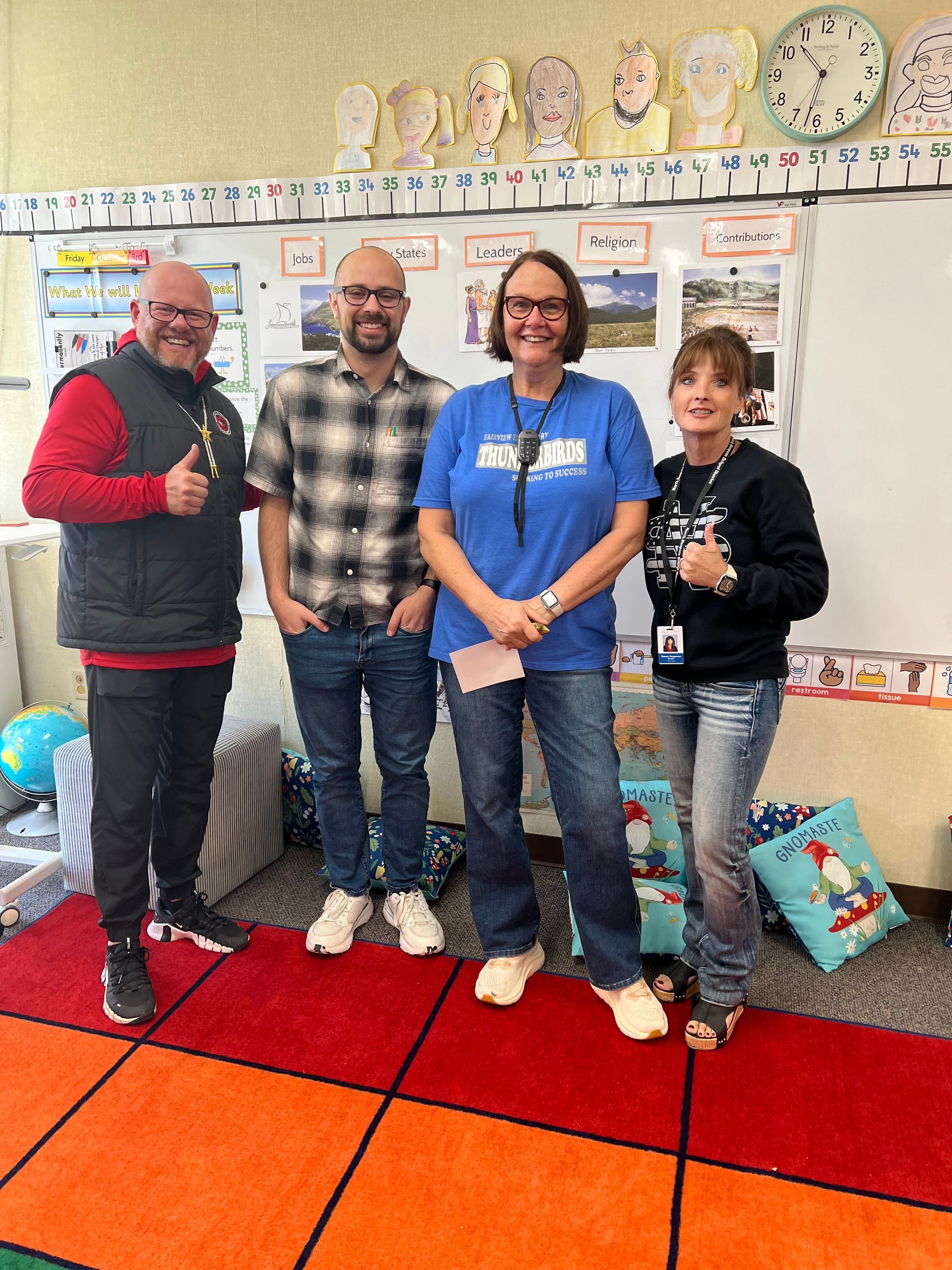Four adults stand in a classroom. The adult on the left gives a thumbs up. They are standing on a colorful rug.