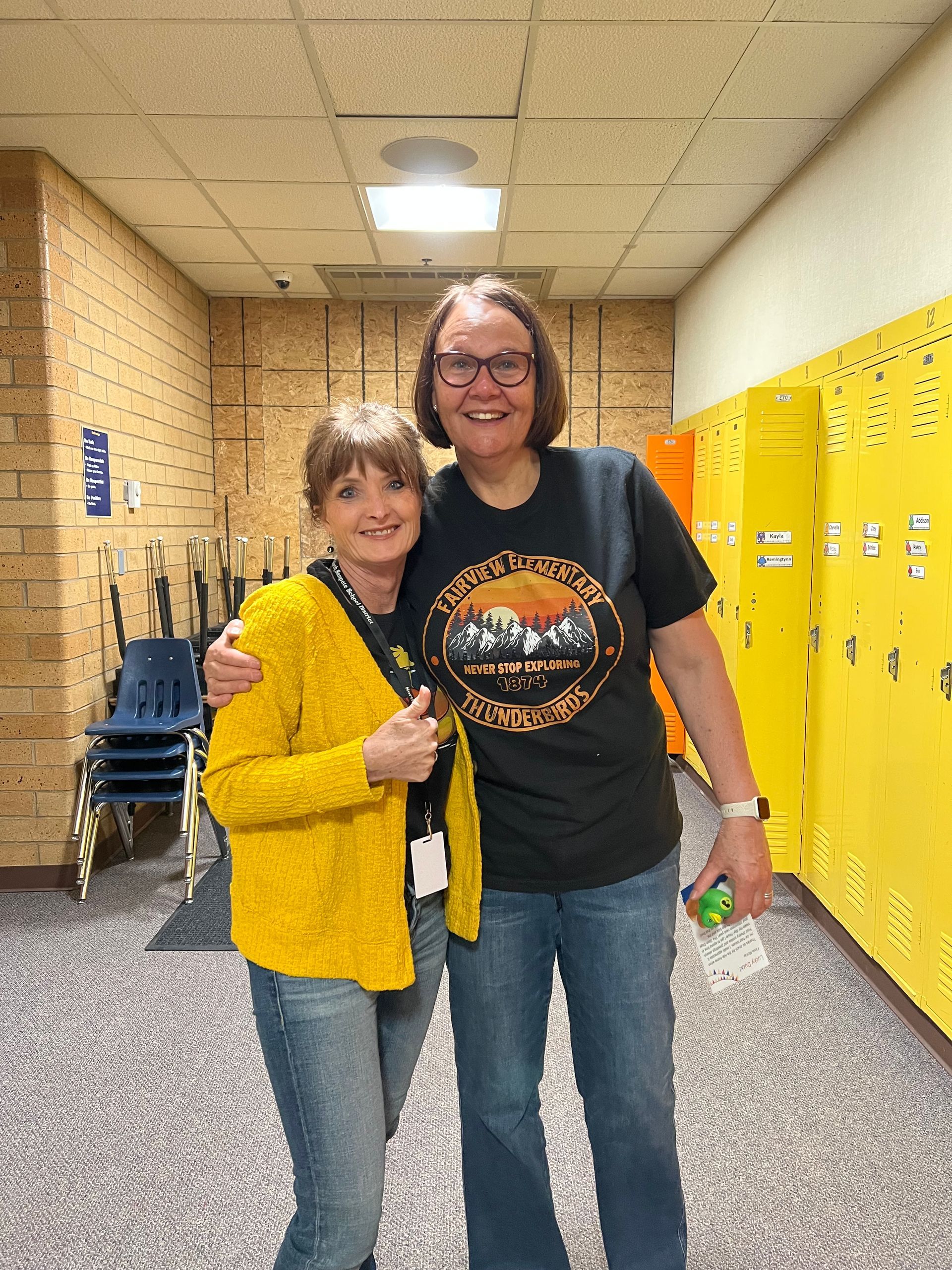 Two women are posing for a picture in a hallway with yellow lockers.