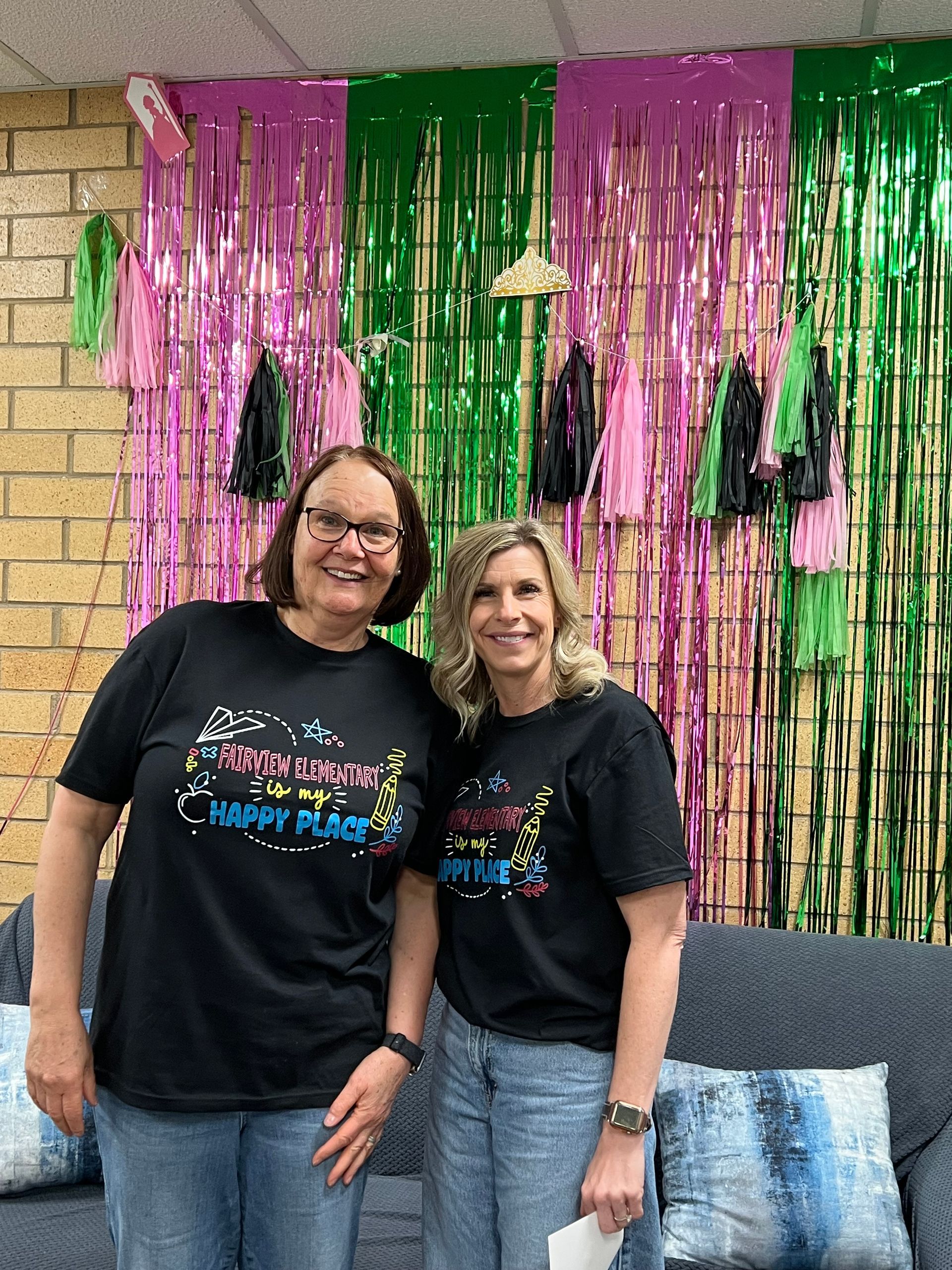 Two people in black T-shirts stand smiling before pink and green streamers on a brick wall.