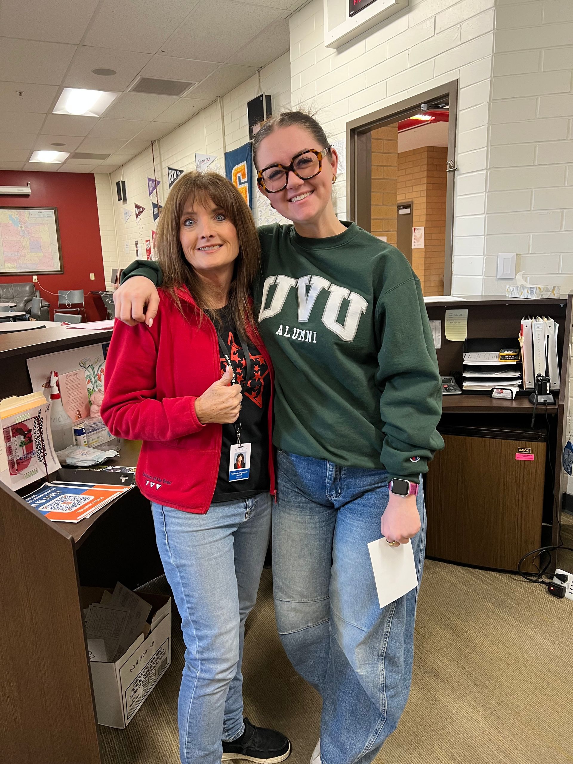 Two women smiling, one in red jacket, the other in green sweatshirt, arms around each other in a school setting.