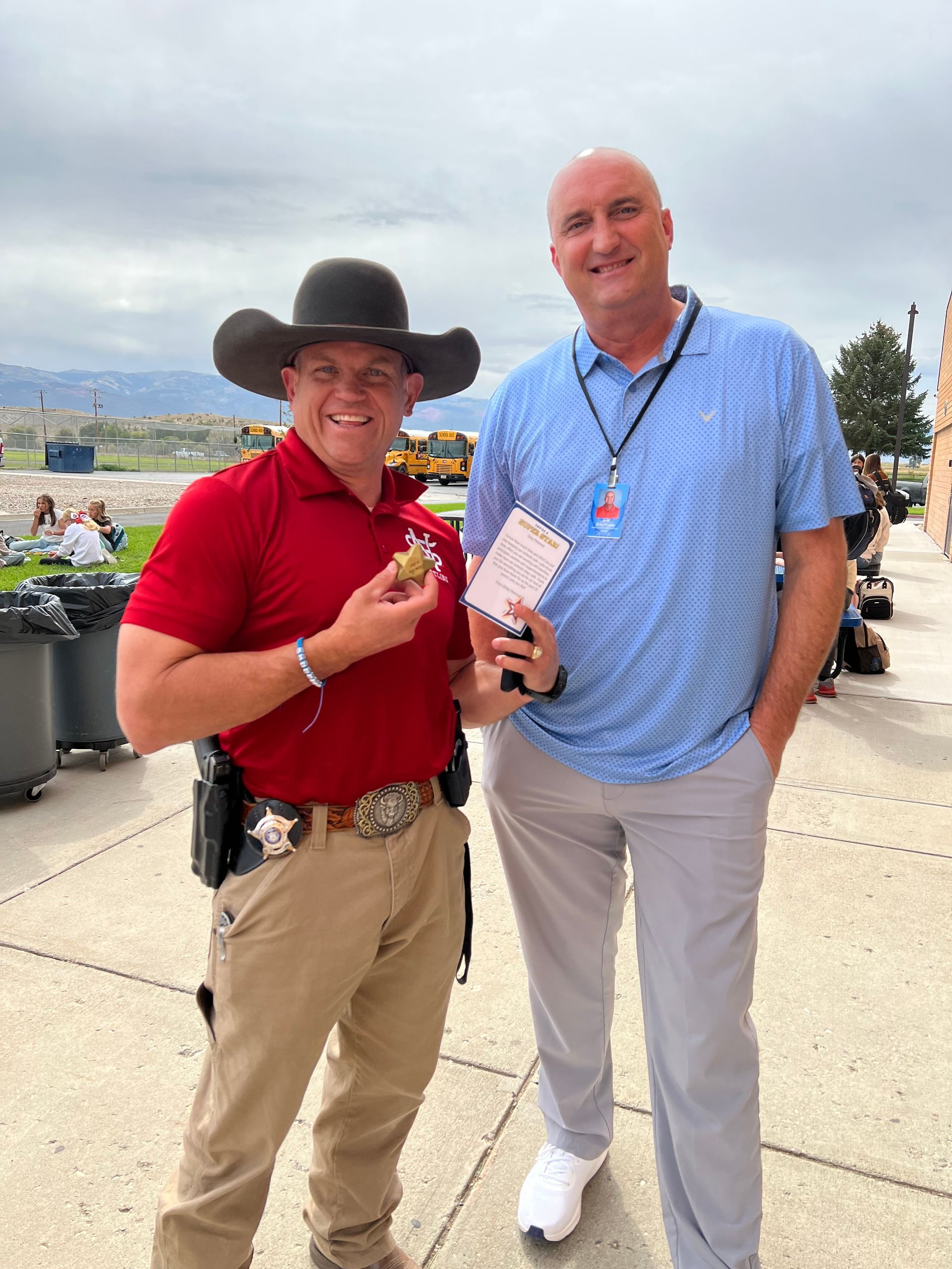 A man in a cowboy hat holds a gold coin with a man in a blue shirt smiling outside.