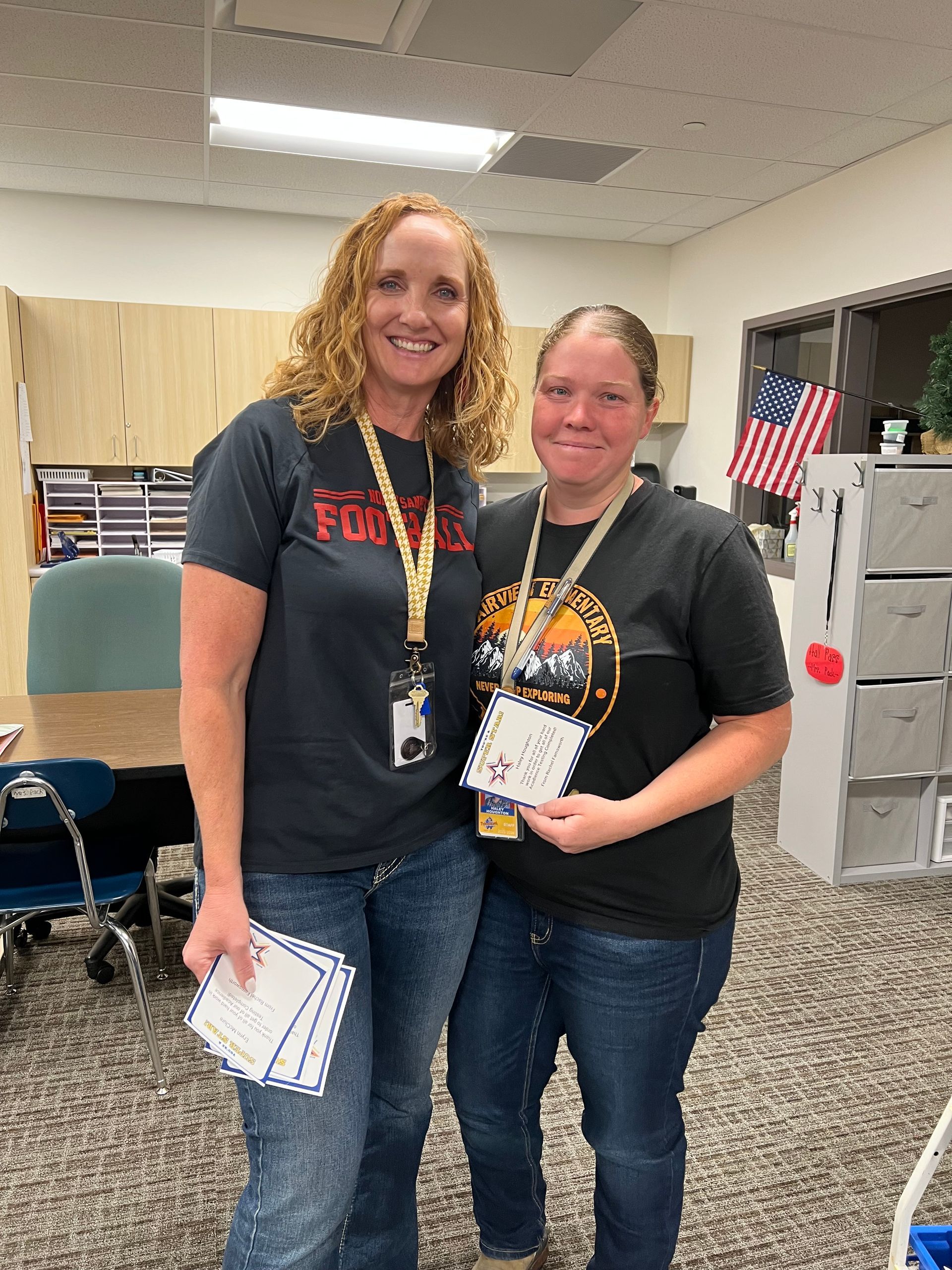 Two women smiling, holding papers, in a classroom. One with curly red hair, wearing black shirt, and the other with brown hair.