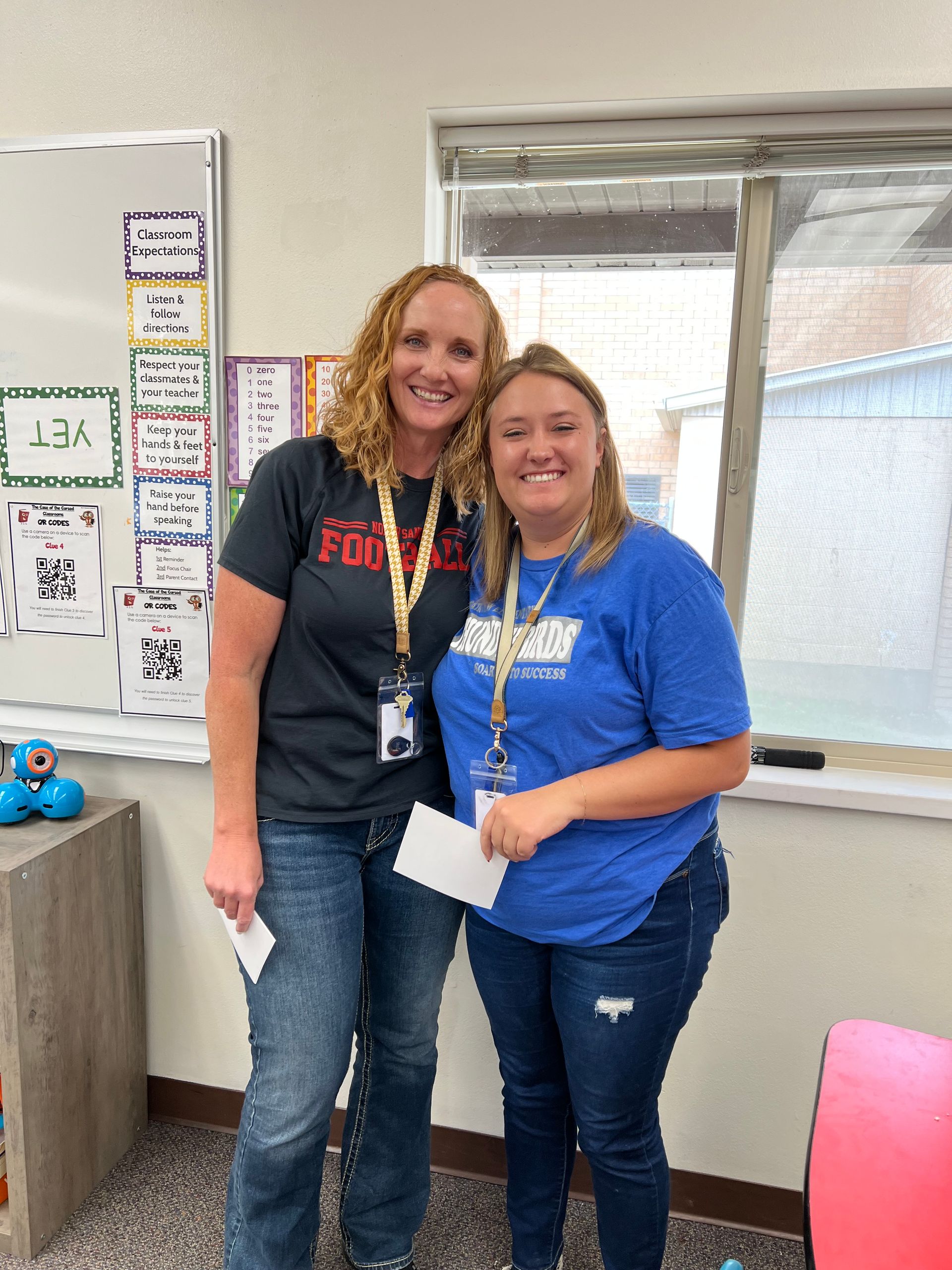 Two women smiling, posing in classroom. One in black shirt, other in blue. Holding papers.