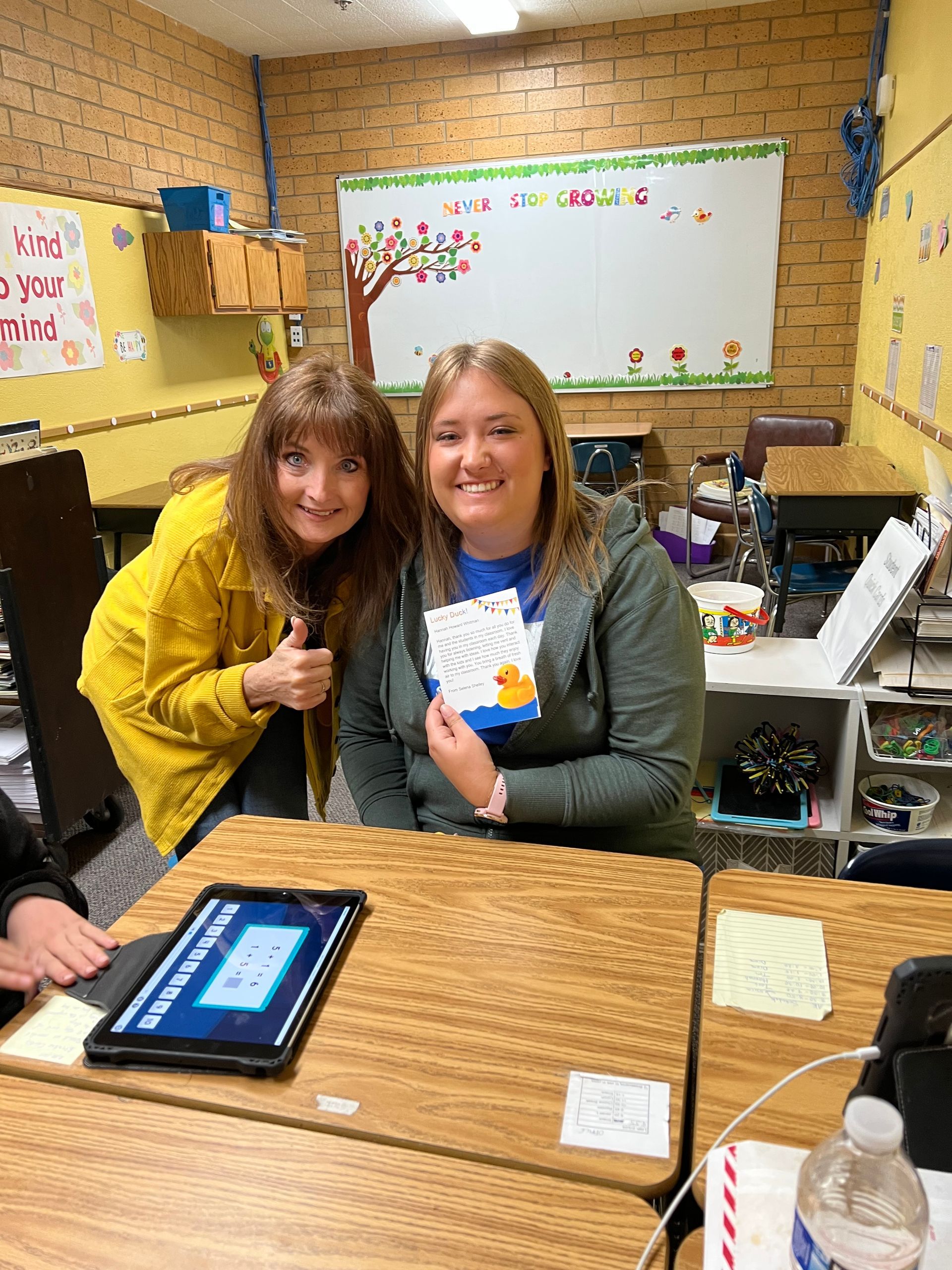 Two women are sitting at a desk in a classroom holding cards.